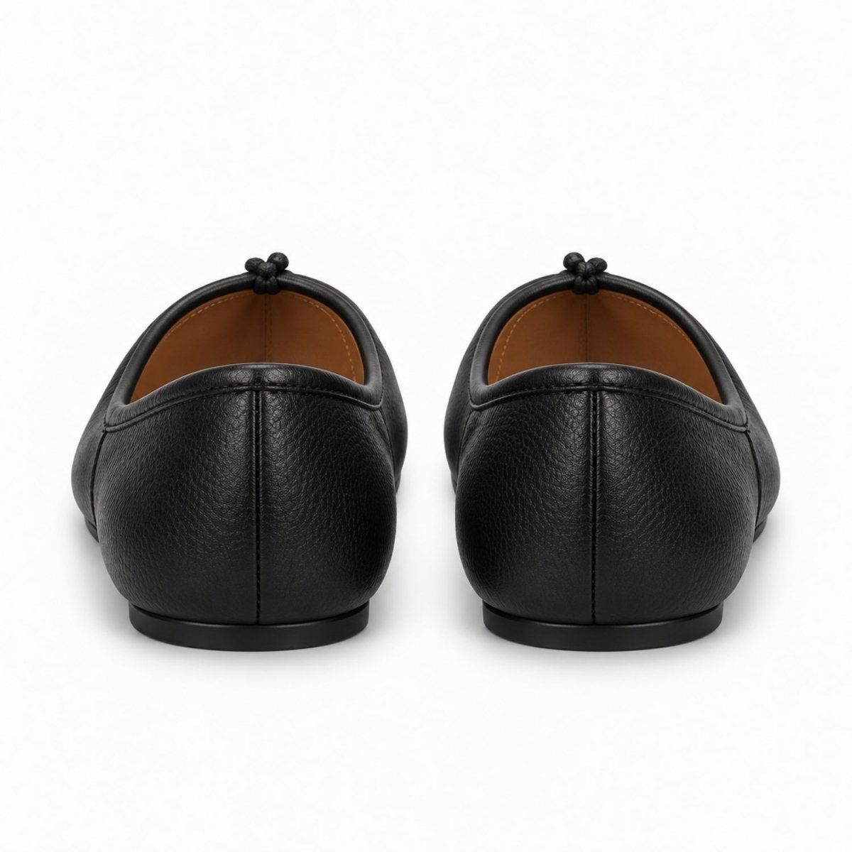 A pair of black pebbled leather ballet flats shown from the back, featuring a small black tassel detail on the back of the shoe and a contrasting tan interior lining, set against a plain white background.