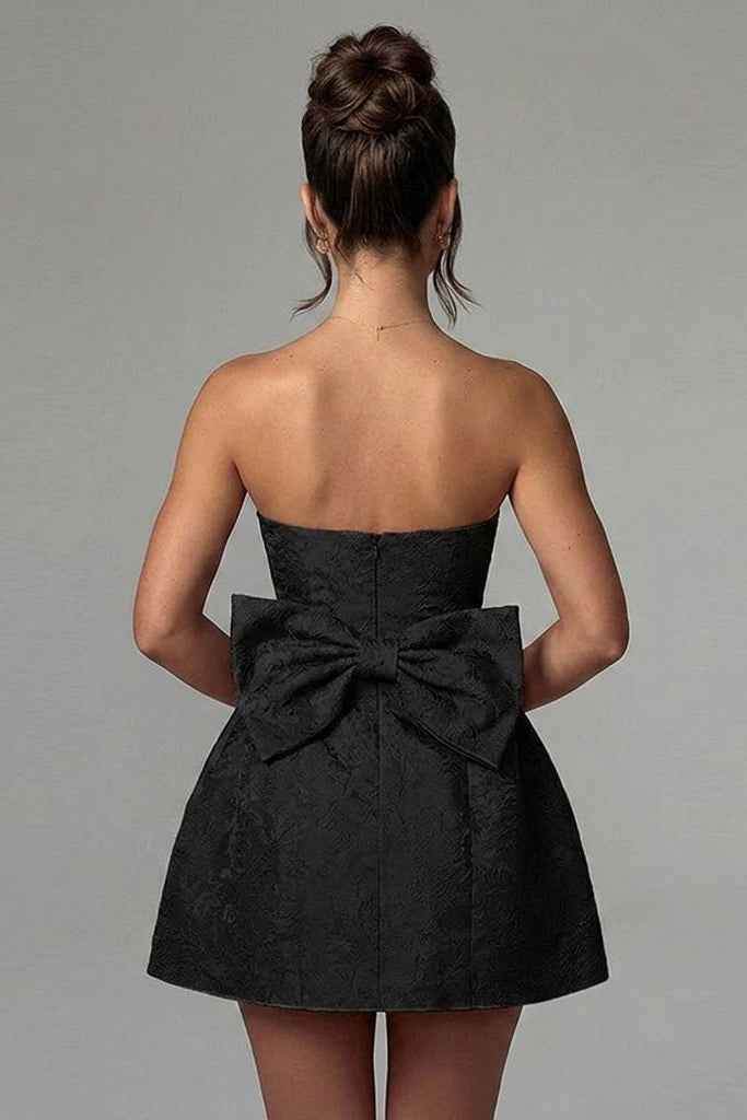 Back view of female model wearing a black mini dress featuring a large statement bow and floral textured fabric by Kouvr.