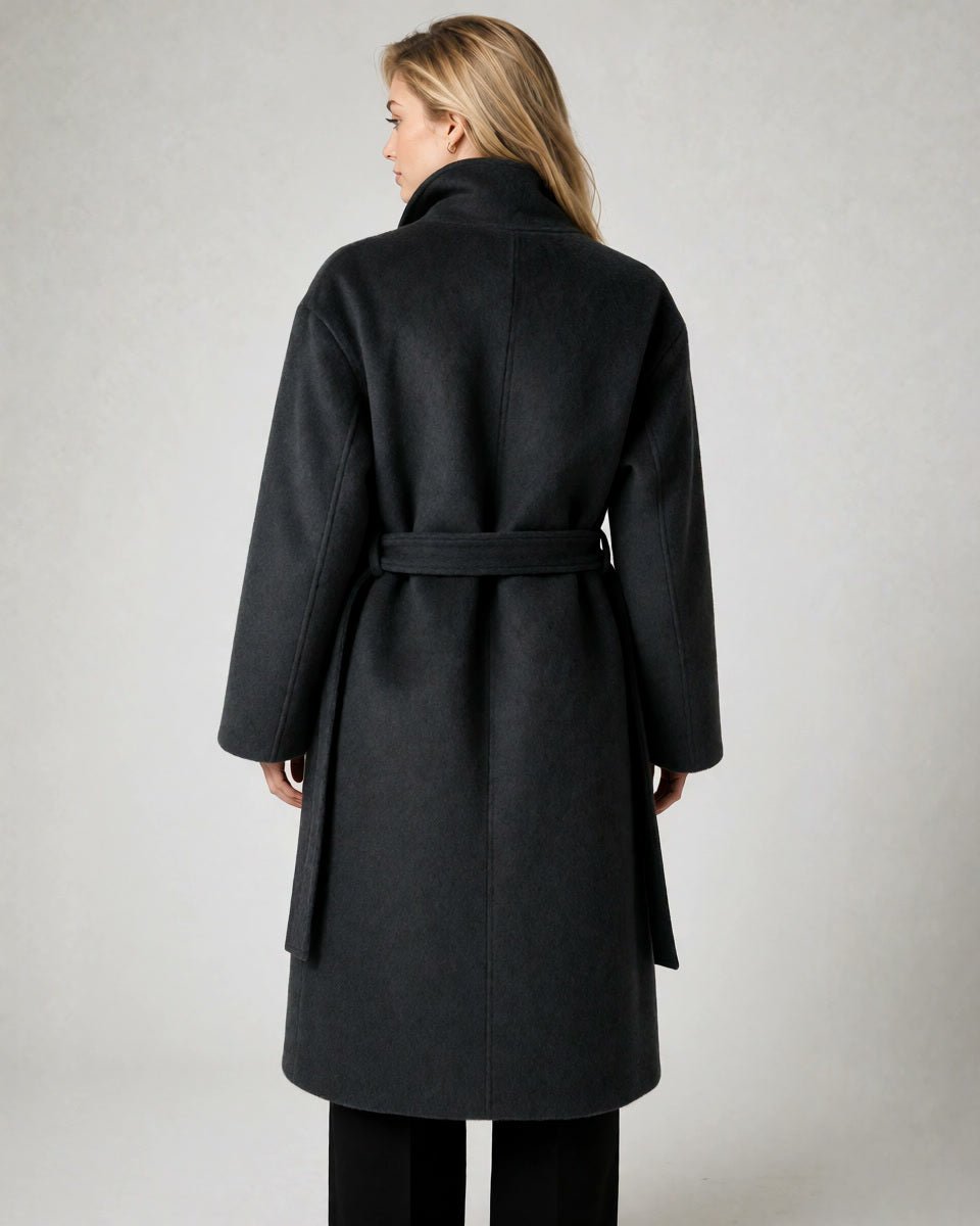 A woman viewed from the back wearing a dark charcoal gray belted coat with a high collar, paired with black pants, against a neutral light gray background.