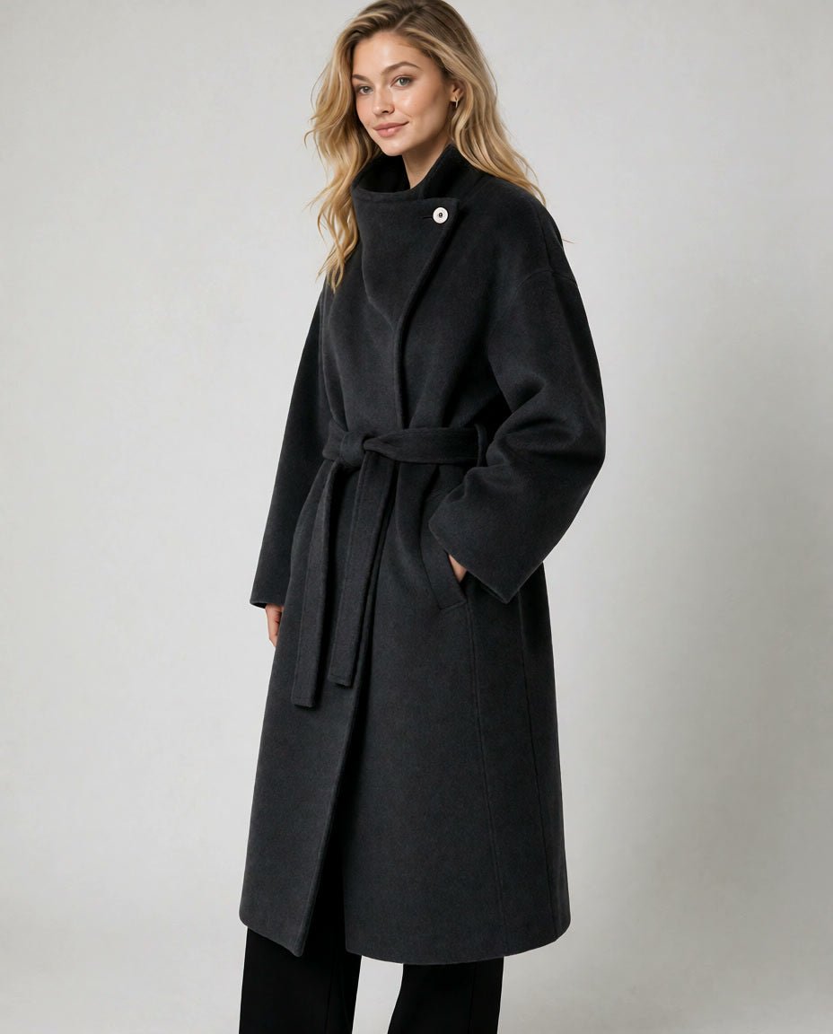 A woman modeling a long black wrap coat with a self-tie belt and a single decorative button at the collar, photographed against a plain light gray studio background.