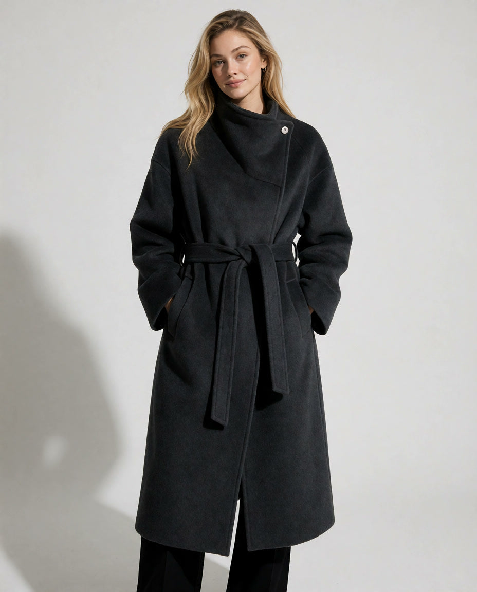A woman modeling a long black wrap coat with a tie belt and asymmetrical button closure, photographed against a plain light gray studio background.