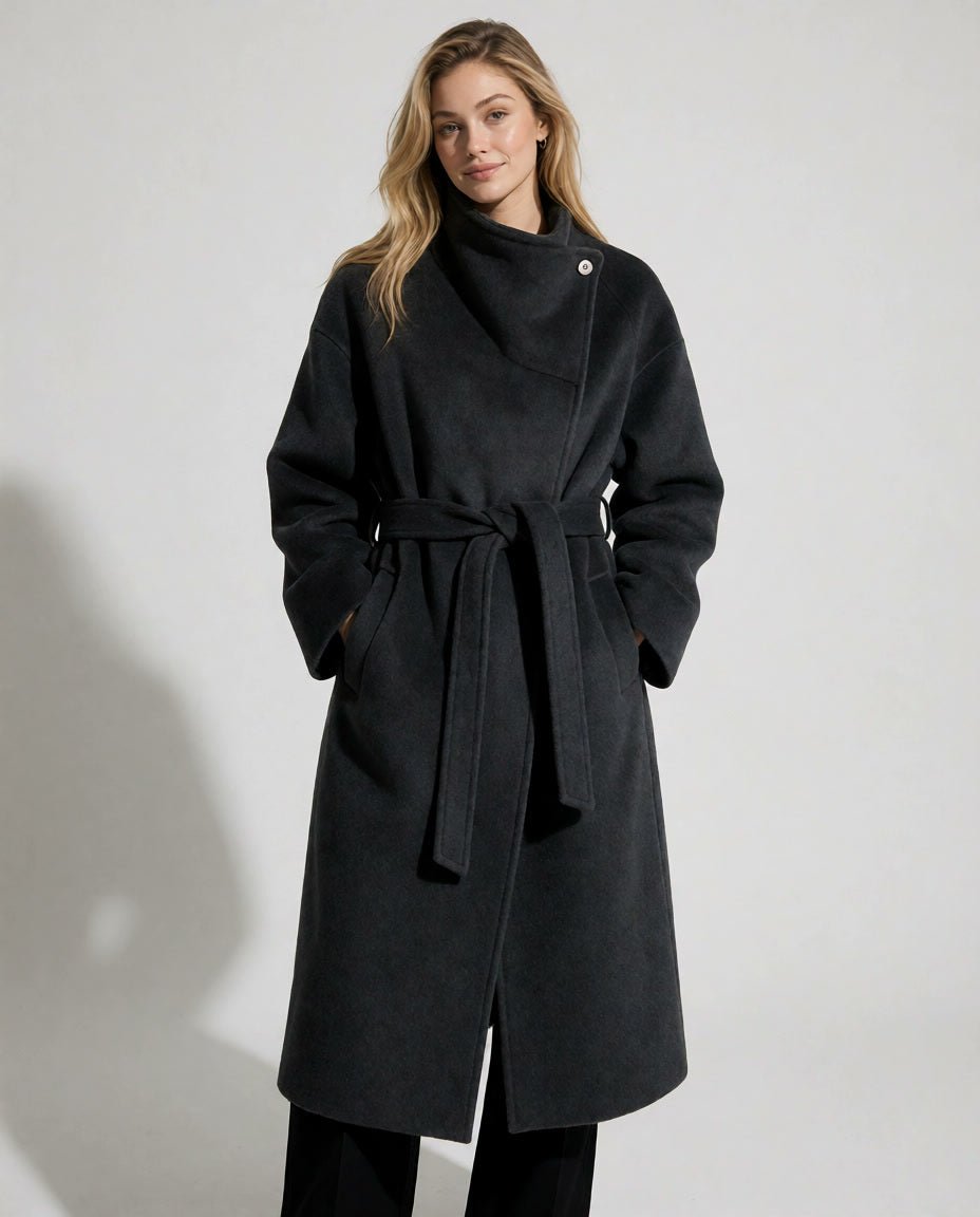 A woman modeling a long black wrap coat with a tie belt and asymmetrical button closure, photographed against a plain light gray studio background.