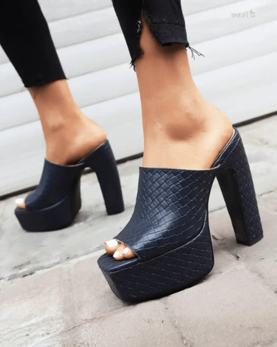 A pair of women's black platform mule heels with a textured crocodile-embossed pattern, shown on a model's feet against a gray concrete and white paneled wall background.