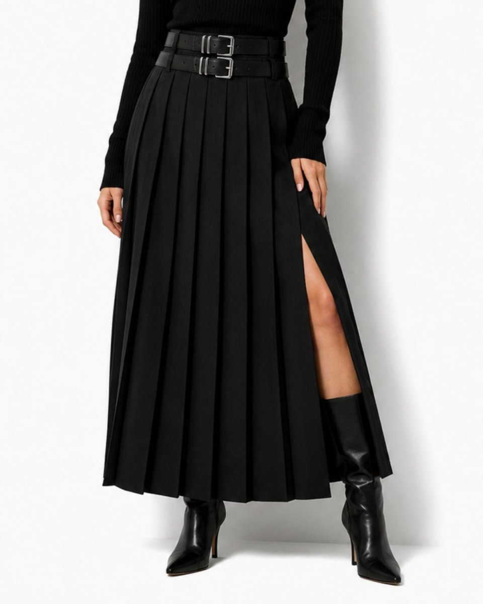 A model wearing a high-waisted black pleated maxi skirt with a double-buckle belt, a black long-sleeve top, and black pointed-toe heeled ankle boots, photographed against a light gray studio background.