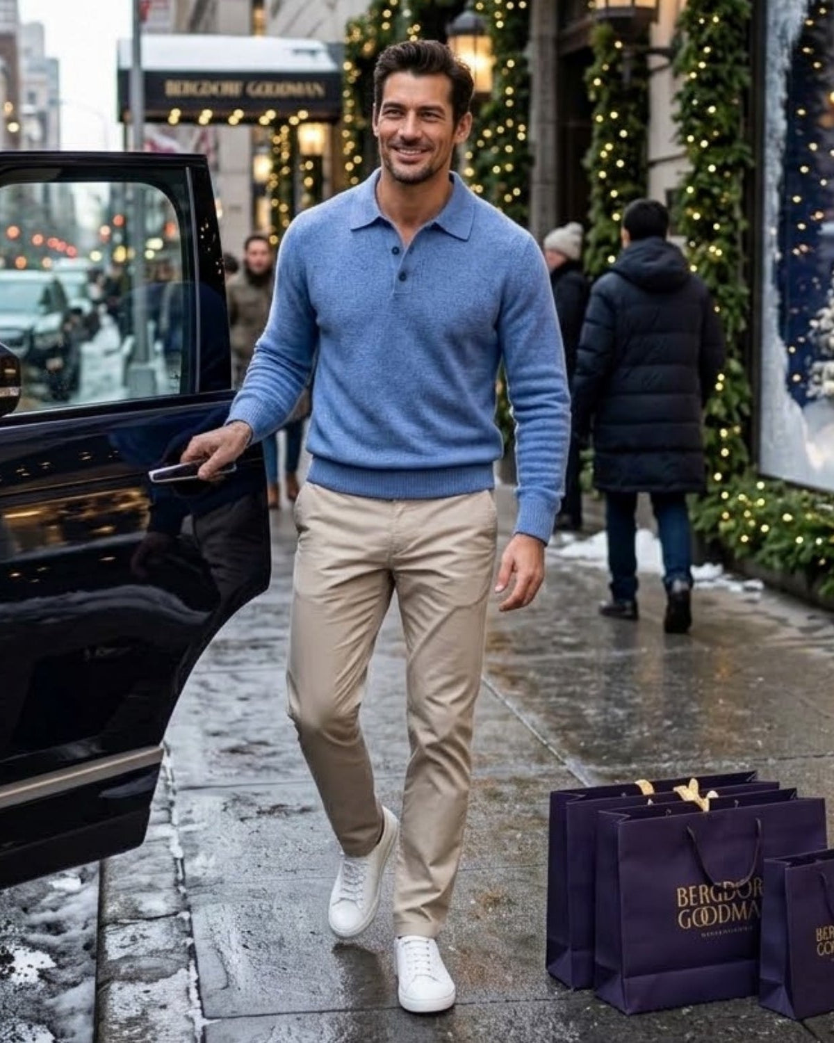 A man in a blue knit polo sweater and beige trousers walks out of a black car on a city sidewalk decorated for Christmas. Several dark purple Belleville Goldman shopping bags are placed on the sidewalk in the foreground.