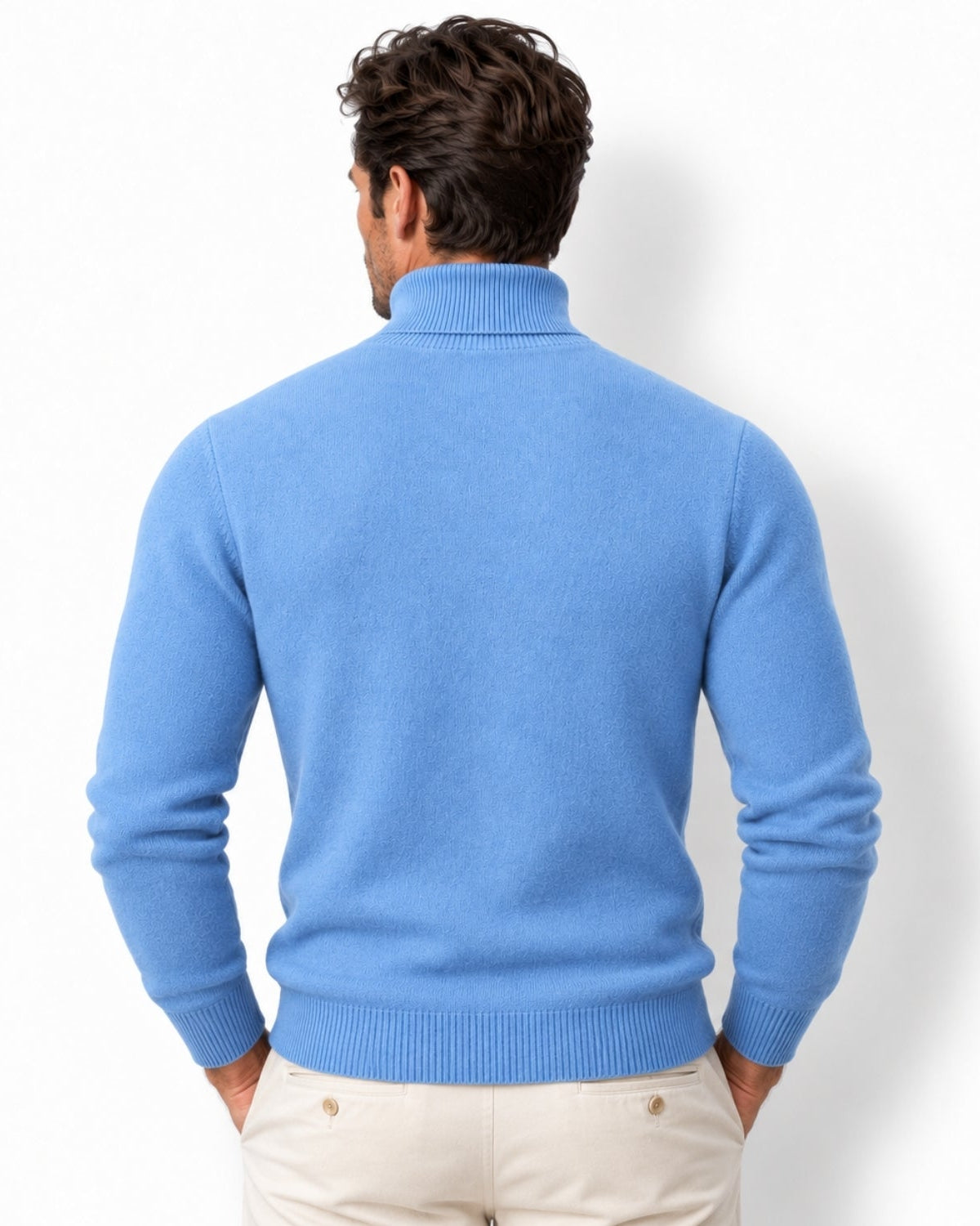 A man viewed from the back wearing a light blue ribbed turtleneck sweater and cream trousers, standing against a plain white studio background.