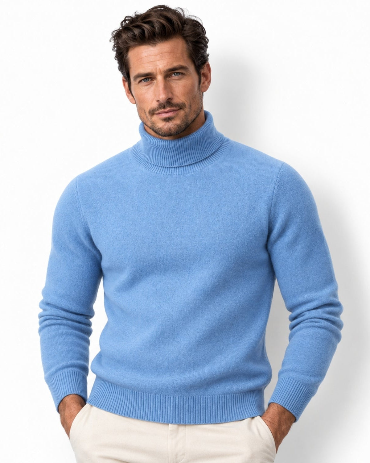 A man wearing a light blue knit turtleneck sweater and cream trousers, standing against a plain white studio background.