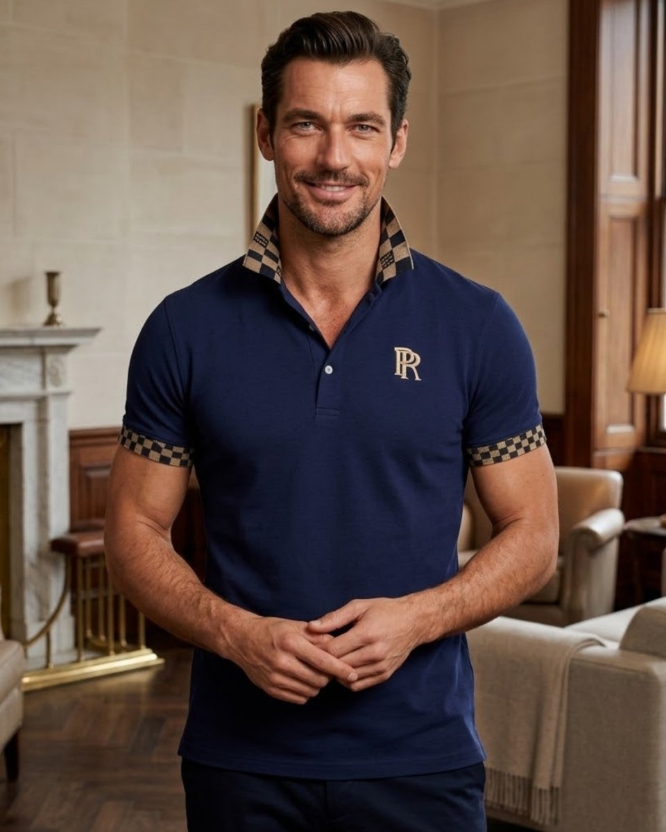 A man wearing a navy blue polo shirt with a gold embroidered 'RR' logo on the chest and a checkered patterned collar and sleeve cuffs, standing in a warm, elegant interior room.