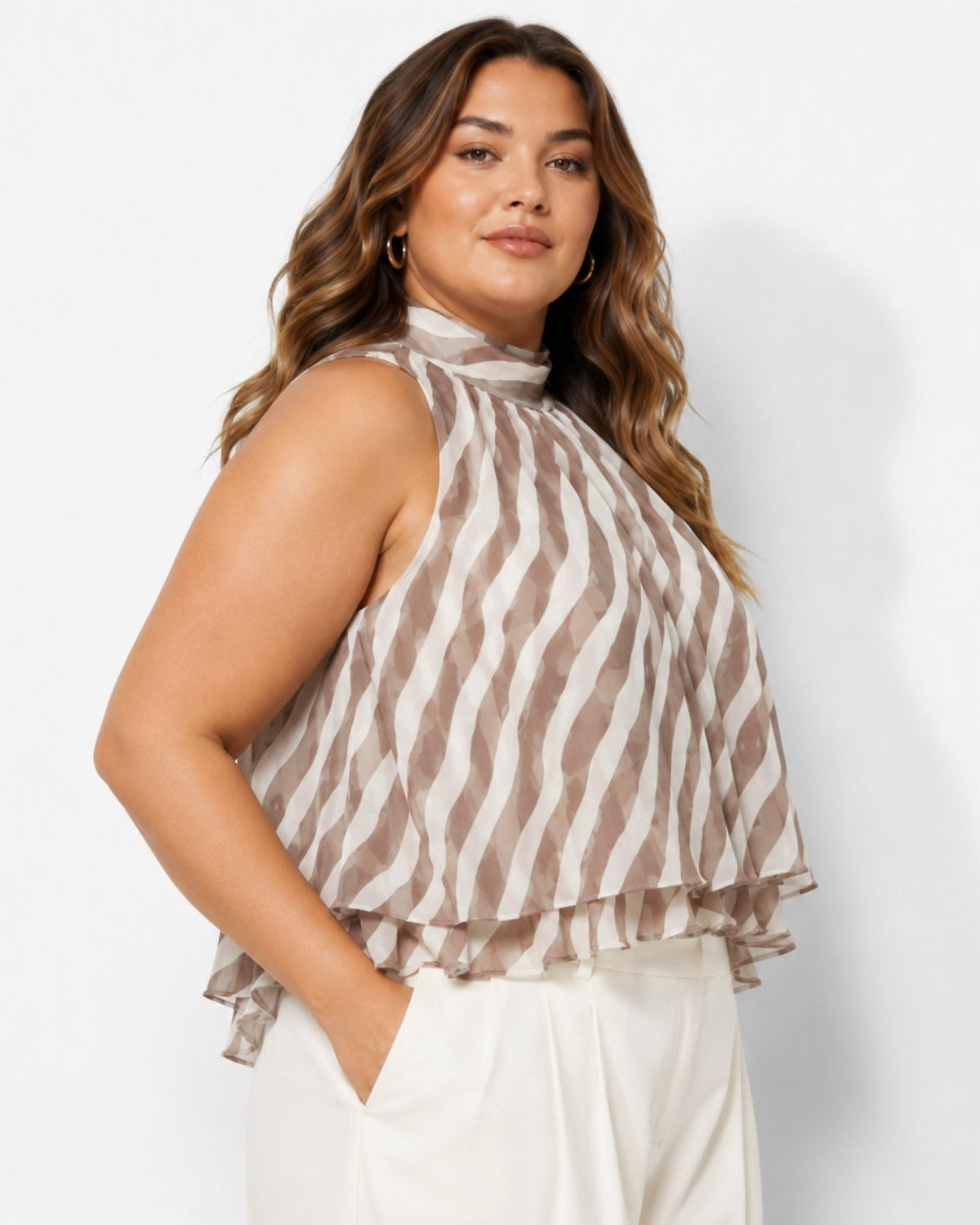 A woman wearing a sleeveless high-neck top with a brown and beige wavy stripe pattern, paired with white pants, photographed against a light gray studio background.