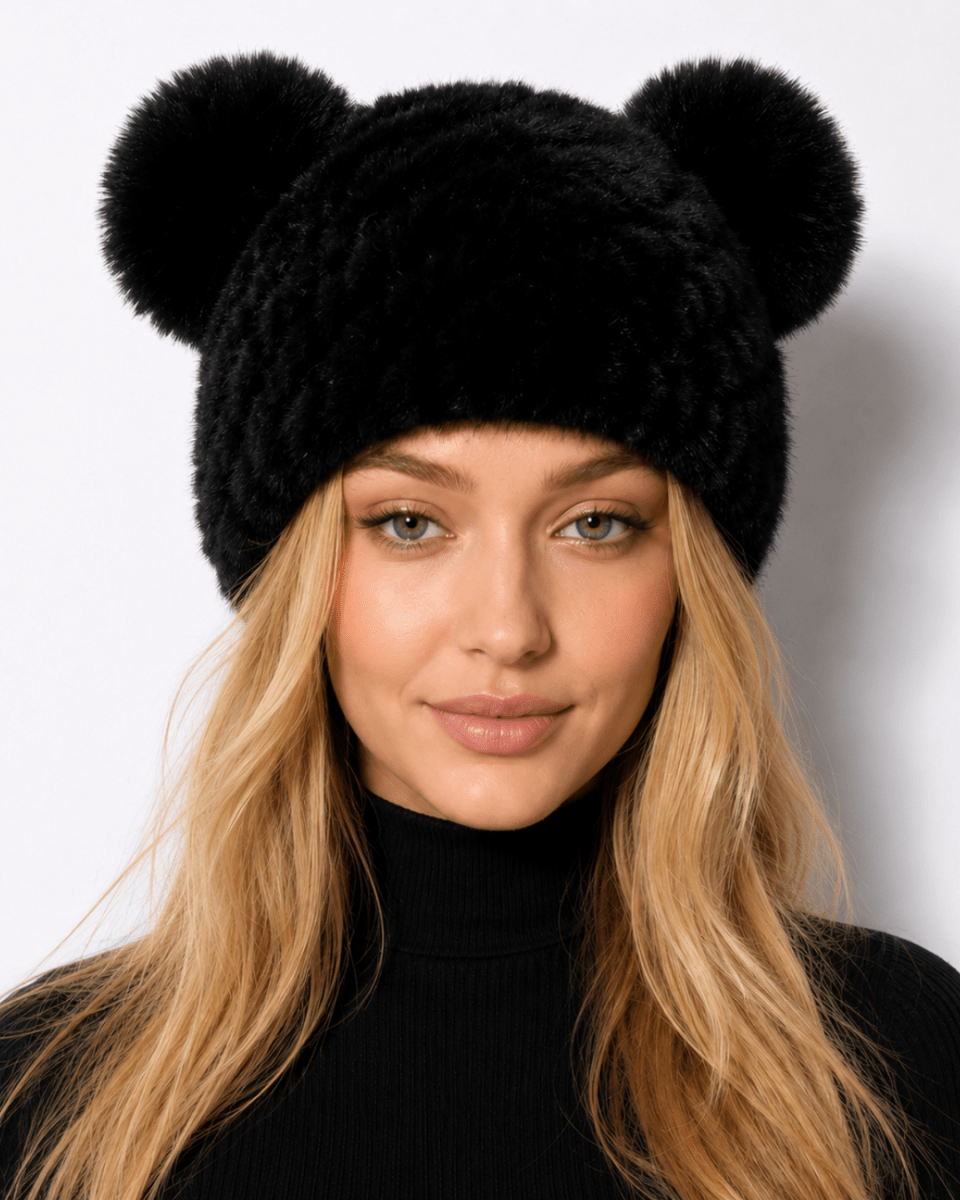 A woman with long blonde hair wearing a black fuzzy beanie hat with two pom-pom ears, photographed against a plain light gray background.