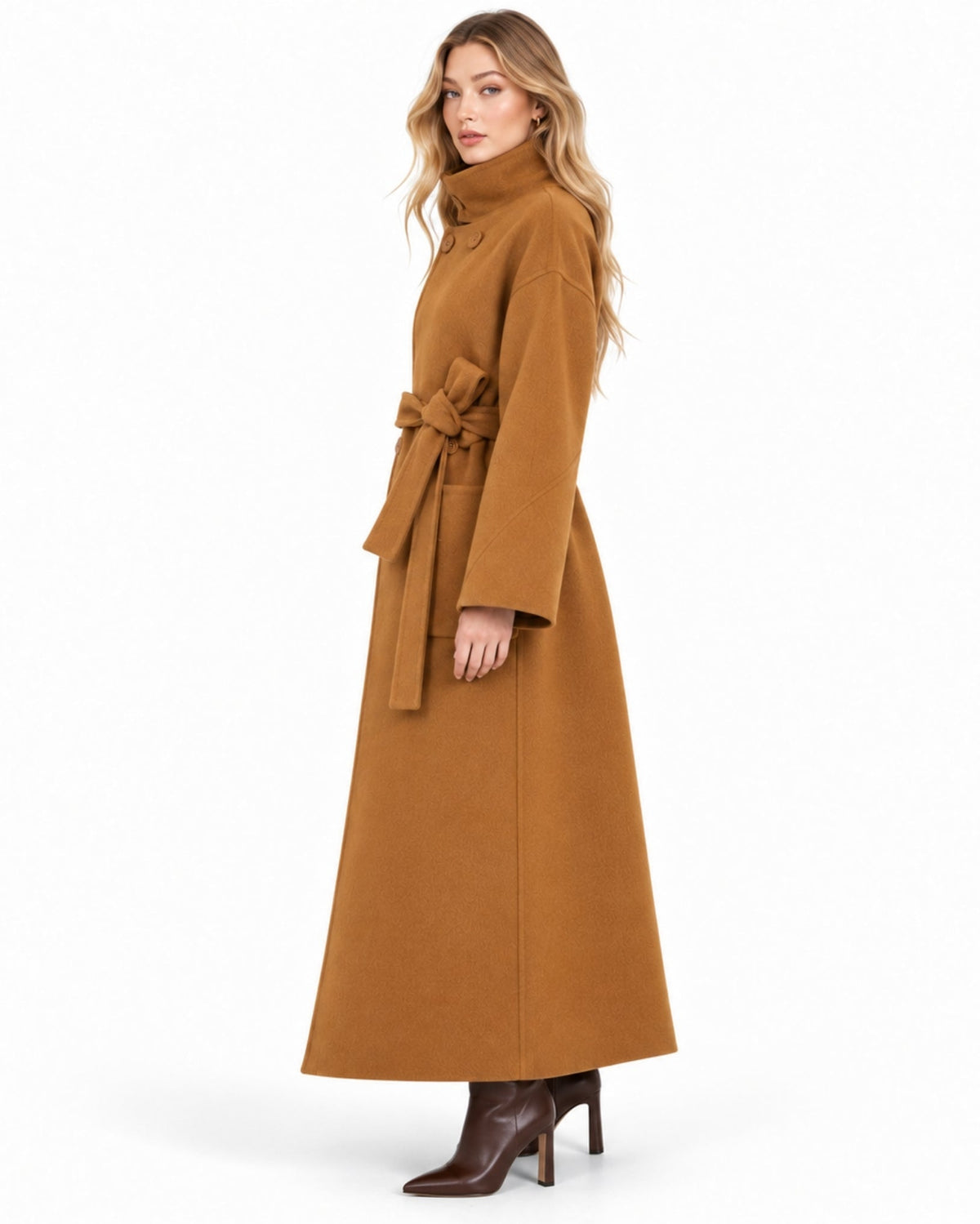 A woman modeling a long, high-neck camel brown coat with a matching waist tie, paired with dark brown heeled ankle boots, against a plain white studio background.