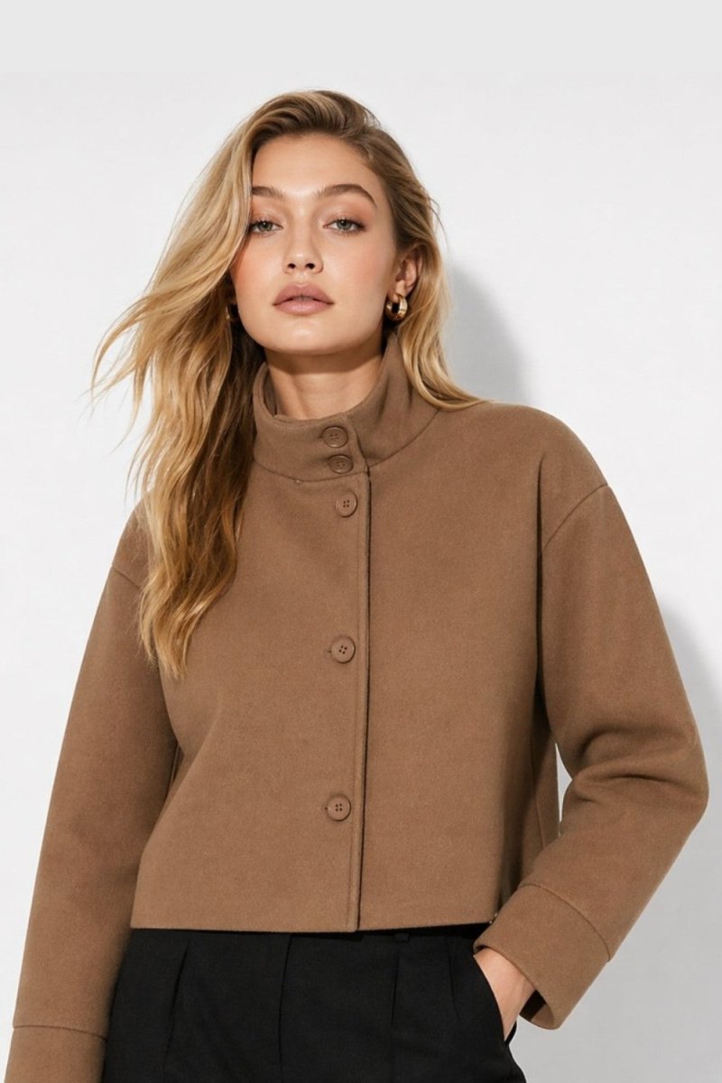 A woman wearing a cropped, long-sleeve camel brown jacket with a high mock neck and three front buttons, paired with black trousers, against a plain light gray background.