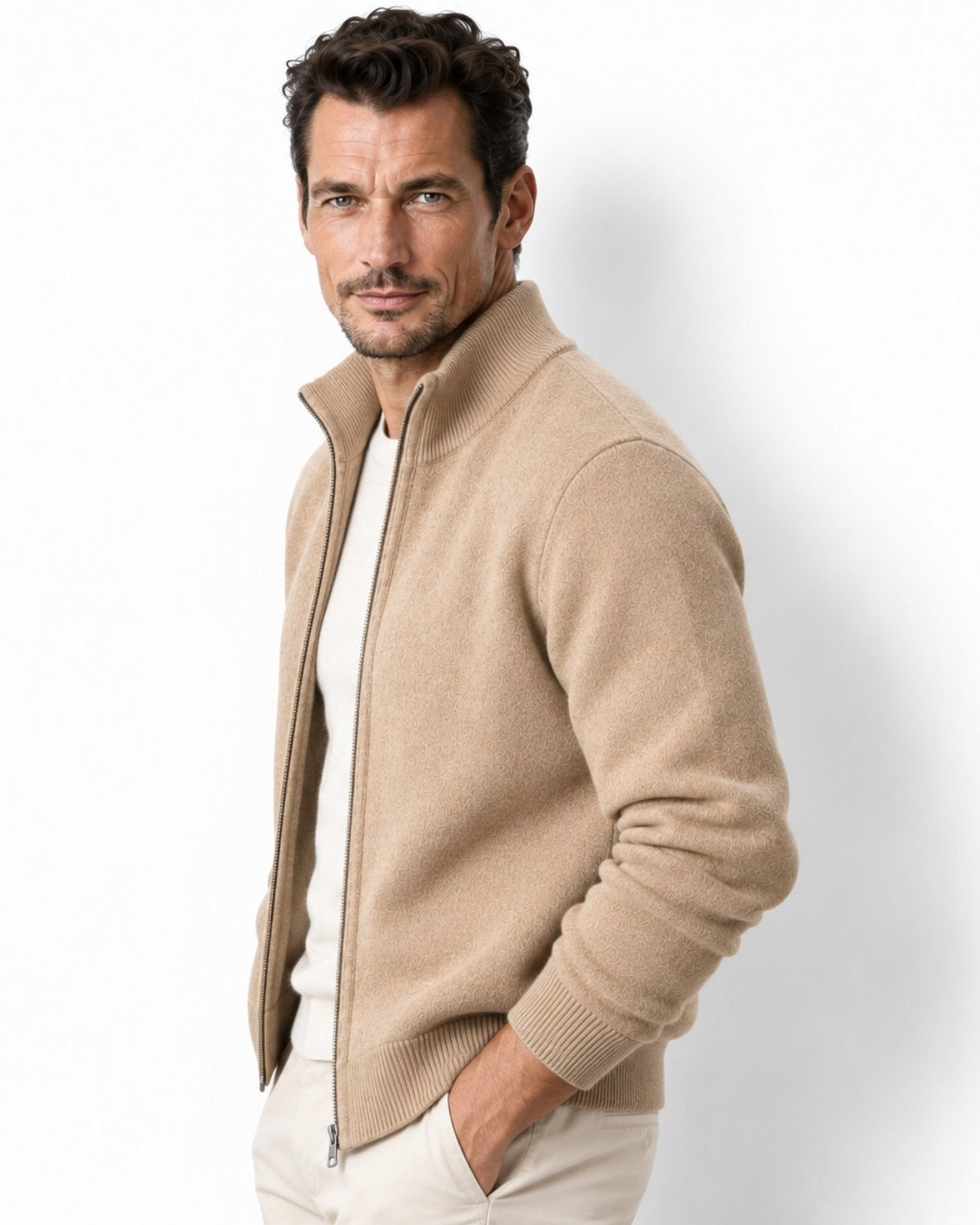 A man wearing a beige knit zip-up sweater over a white shirt and light trousers, photographed against a clean white studio background.