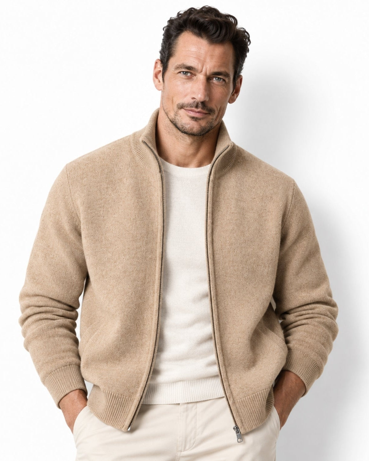 A man wearing a camel knit zip-front bomber-style cardigan over a cream sweater and light trousers, standing against a plain white studio background.