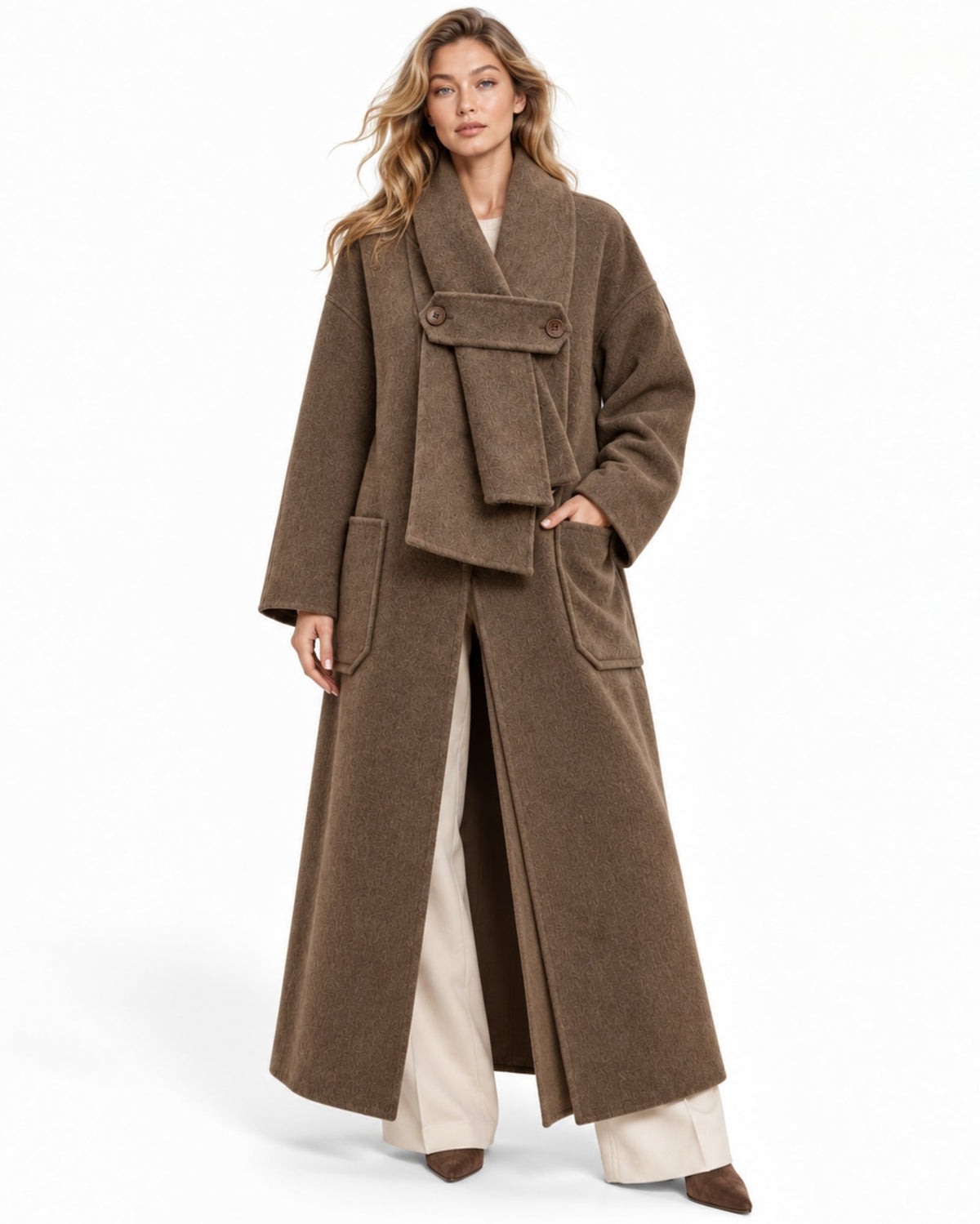 A woman modeling a long brown wrap-style coat with wide lapels, large front patch pockets, and a draped waist detail, paired with cream wide-leg pants and brown pointed shoes against a plain white studio background.