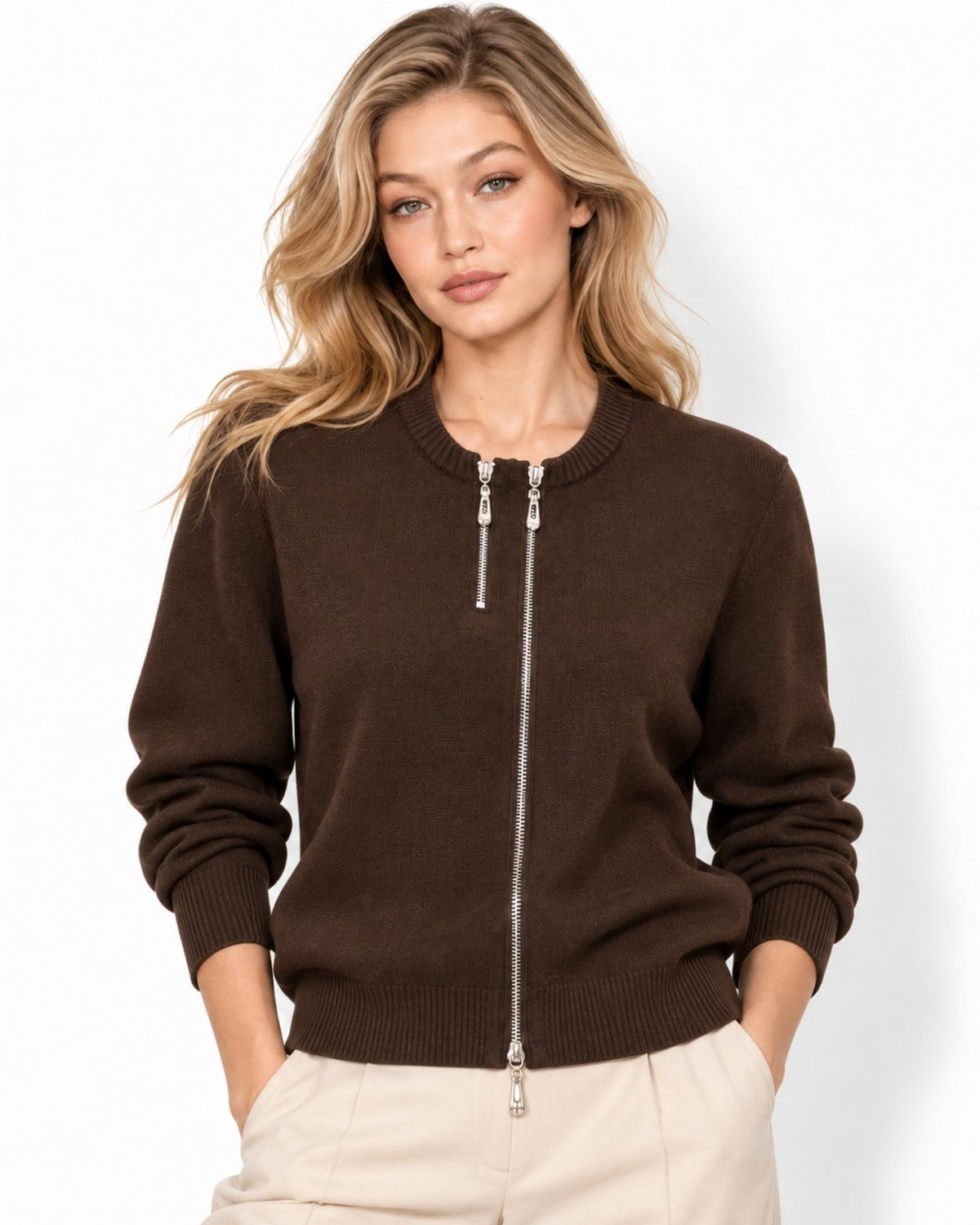 A woman wearing a dark brown zip-up knit sweater with silver-tone zipper pulls and drawstrings, paired with light beige pants, against a plain white studio background.