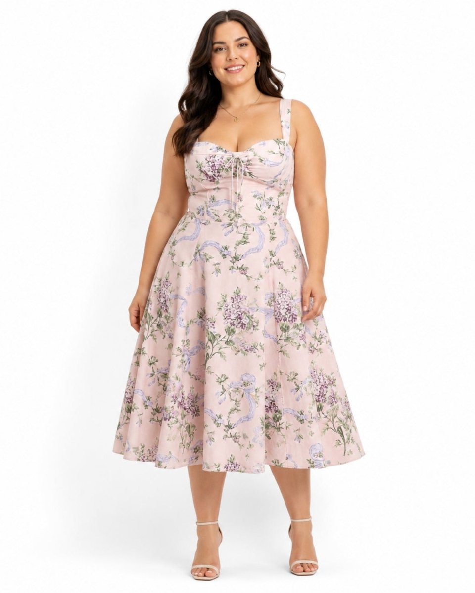 A woman modeling a sleeveless pink midi dress with a purple and green floral print, photographed against a plain white studio background.