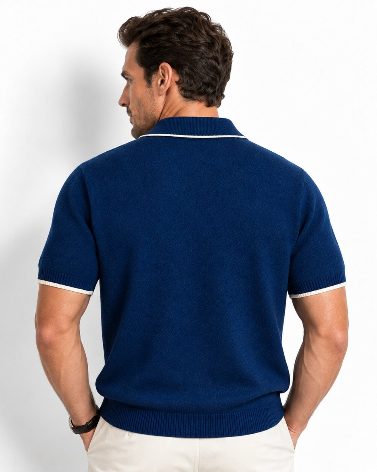 A man shown from the back wearing a navy blue short-sleeve knit polo shirt with white contrast trim at the collar and sleeve cuffs, paired with off-white trousers, against a plain white studio background.