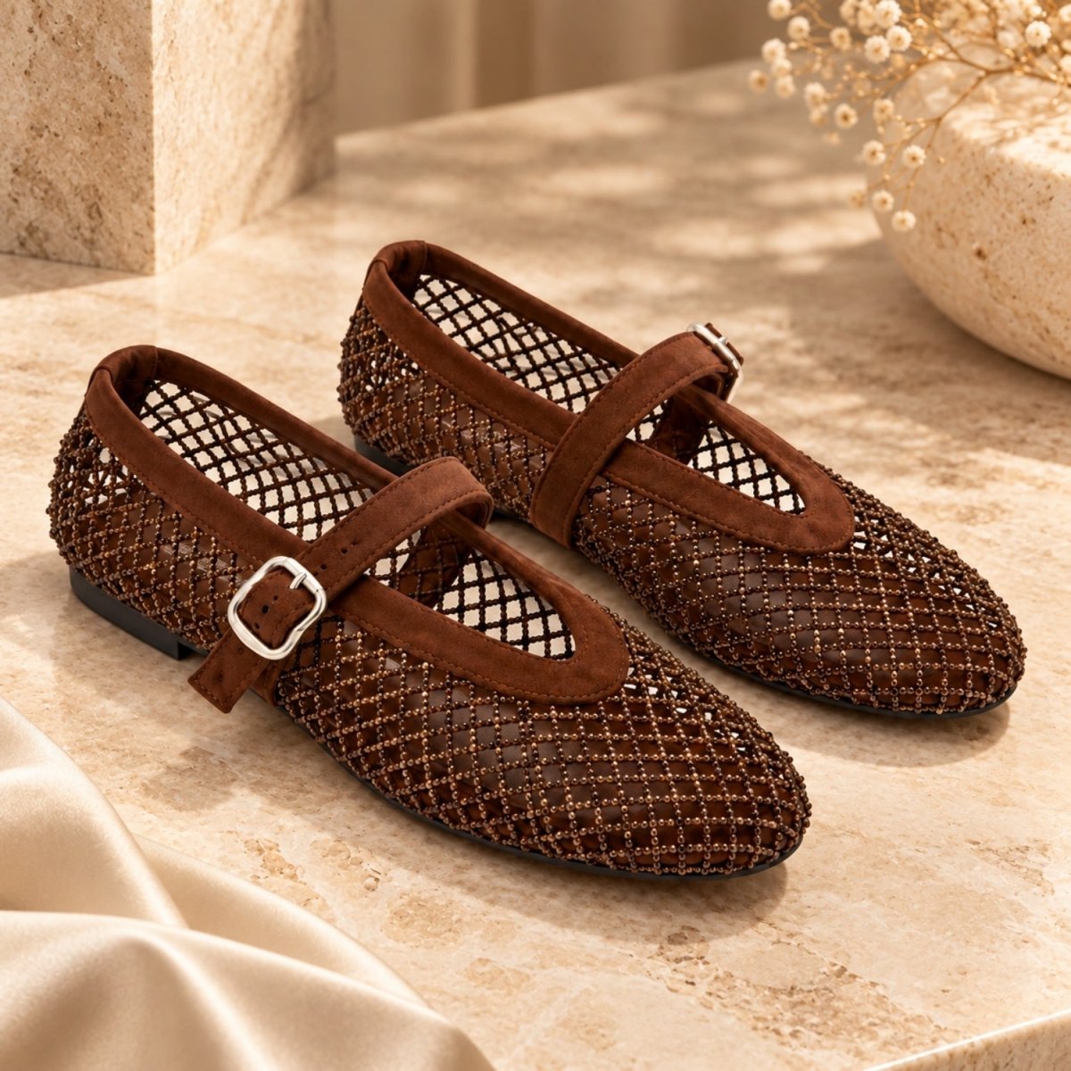 A pair of women's Mary Jane flats in brown, featuring a textured openwork mesh upper, a medium brown strap, and a silver buckle, displayed on a beige stone surface.