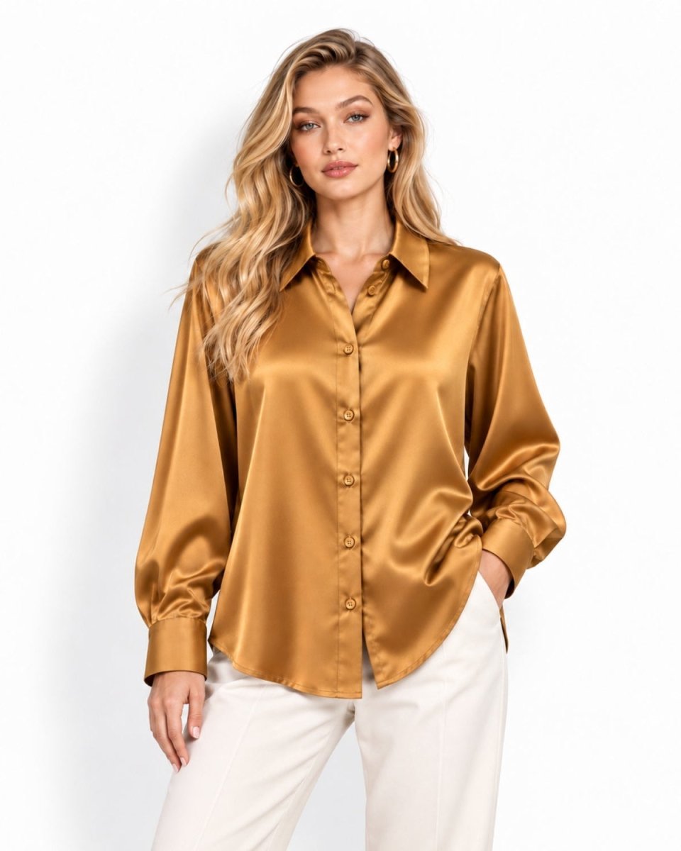 A woman modeling a long-sleeve silk button-up blouse in a warm camel-gold color, paired with white pants, against a clean white studio background.