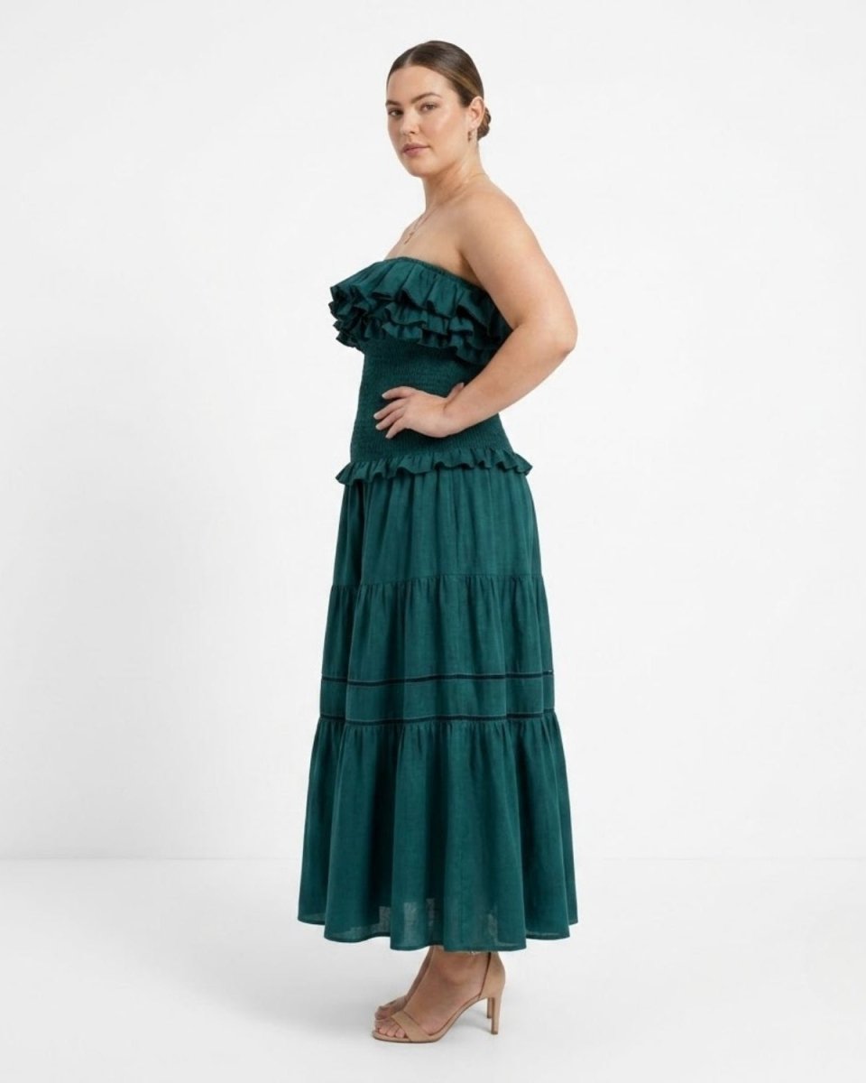 A woman modeling a strapless dark green tiered maxi dress with ruffle details, paired with beige high-heeled sandals, against a plain white studio background.