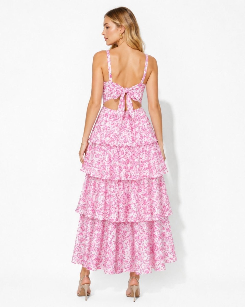 A woman modeling a pink and white floral tiered maxi dress from the back, featuring thin straps, a cutout back with a bow, and a long ruffled skirt, set against a plain white studio background.
