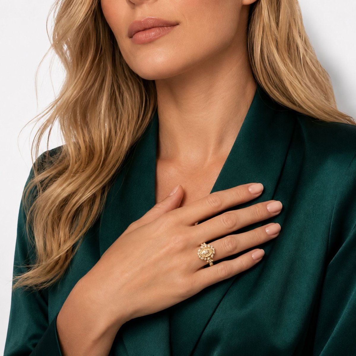 A close-up lifestyle shot of a woman wearing a detailed gold ring on her finger, with her hand resting on her chest. She is wearing a dark green satin robe against a clean white background.