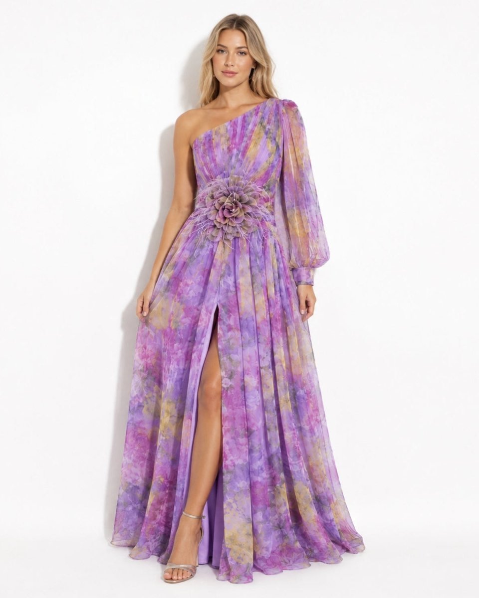 A woman models a one-shoulder floor-length gown with a purple, lavender, and pink tie-dye pattern, featuring a high thigh slit and a large floral embellishment at the waist.