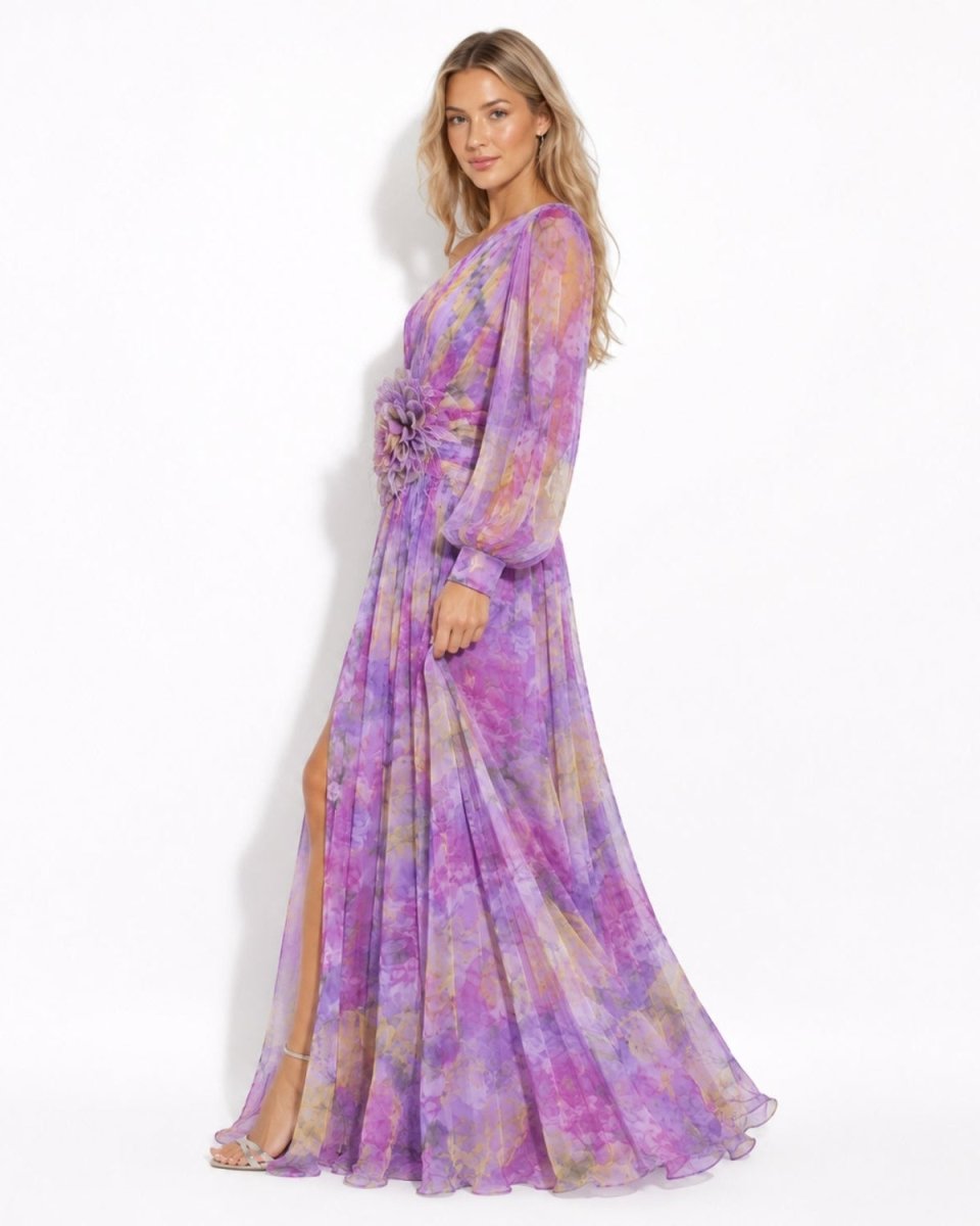 A woman modeling a long-sleeved, floor-length purple and lavender tie-dye maxi dress with a matching fabric flower detail at the waist, set against a plain white studio background.