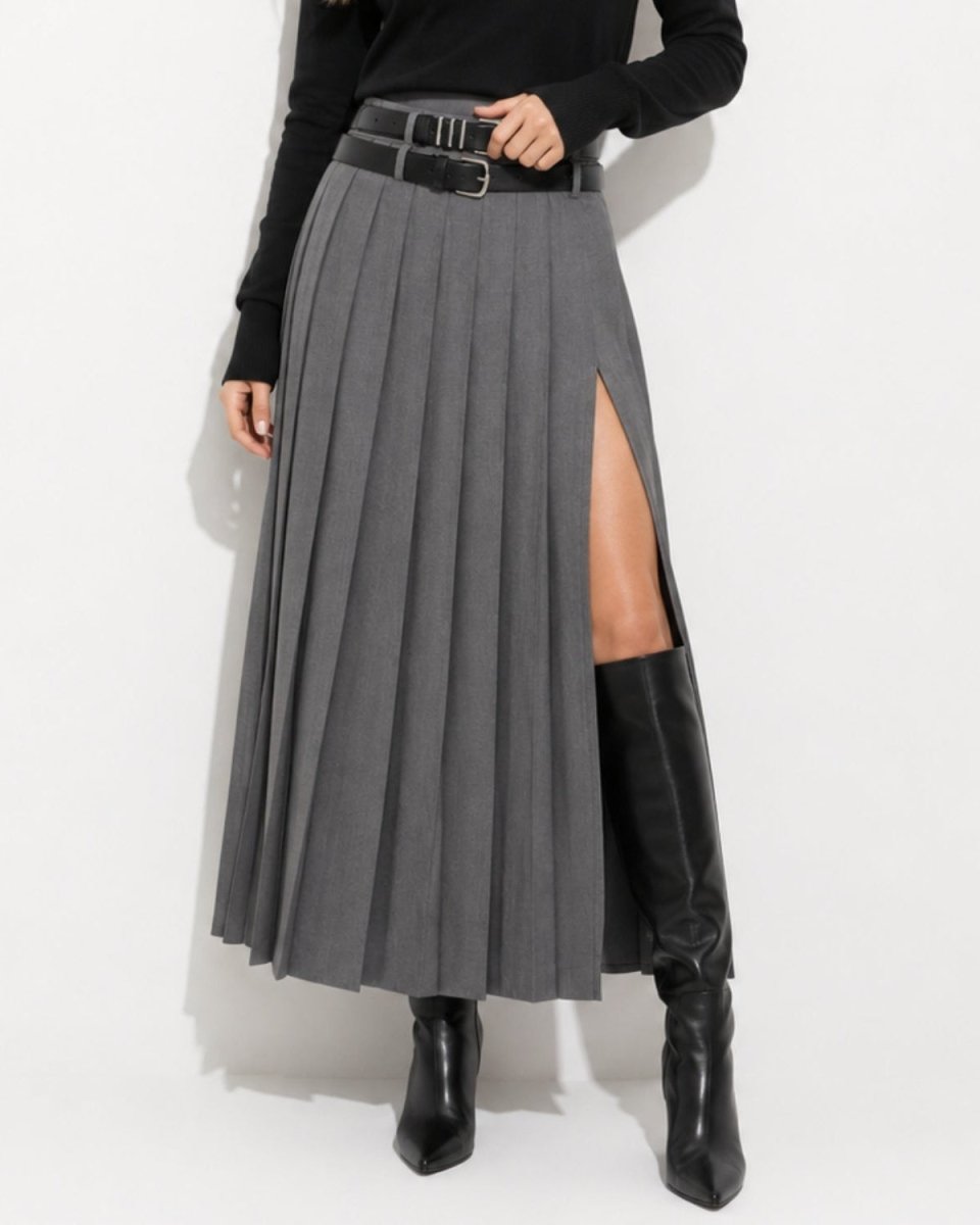 A model wearing a high-waisted, pleated gray midi skirt with a thigh-high side slit, styled with a black belt, a black long-sleeve top, and black knee-high boots against a white studio background.