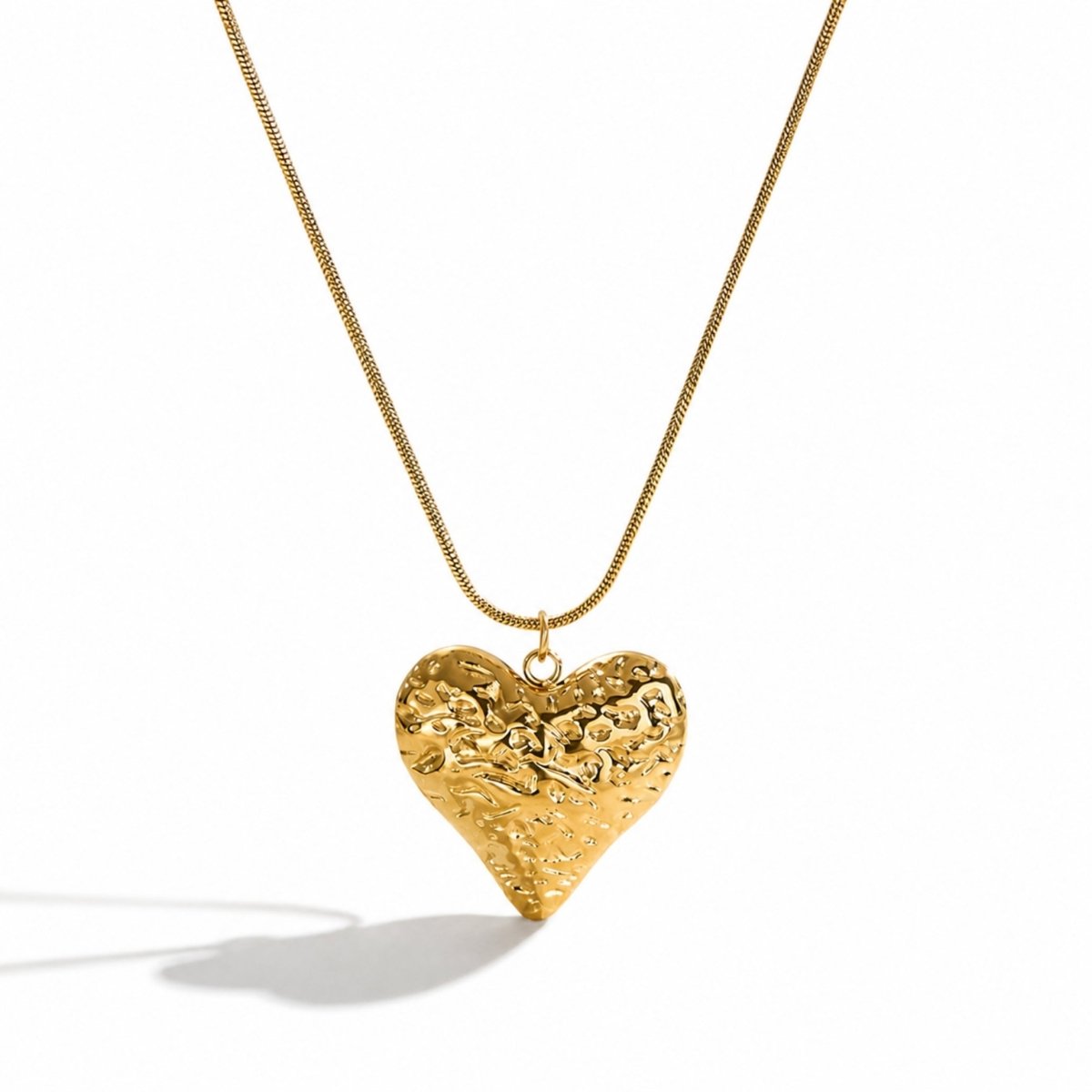 Close up of a shiny polished gold textured heart pendant necklace on a delicate luxury snake chain casting a shadow on a white background
