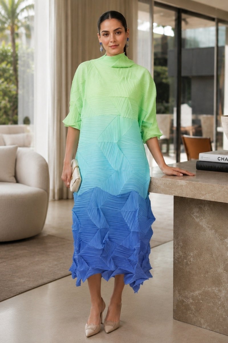 A woman models a three-quarter sleeve midi dress with a vibrant ombre gradient transitioning from lime green at the top, through light blue, to royal blue at the bottom. The dress features a high mock neck and a textured, pleated ruffled hem.