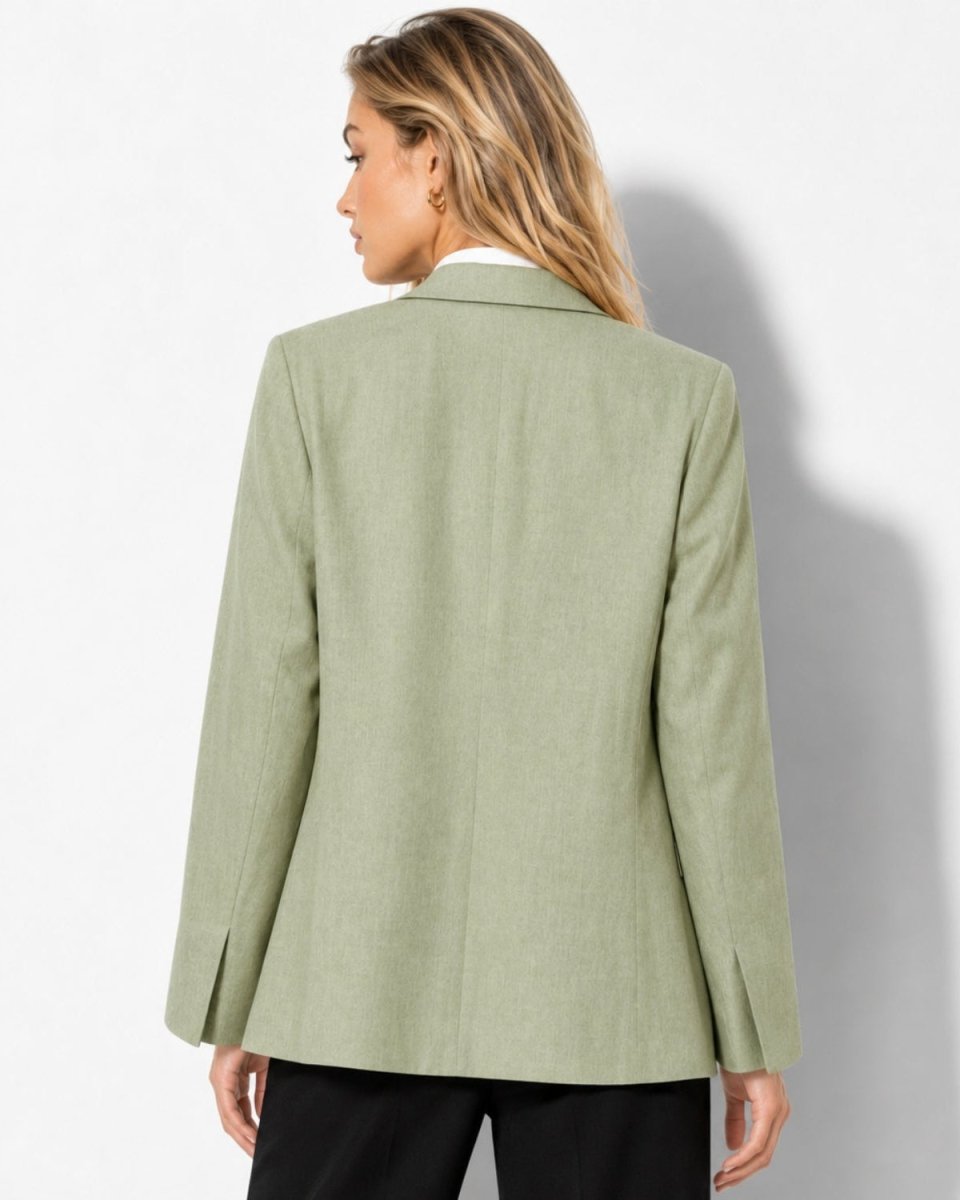 A woman shown from the back wearing a light sage green blazer over a white collared shirt and black trousers, standing against a white wall with a soft shadow.