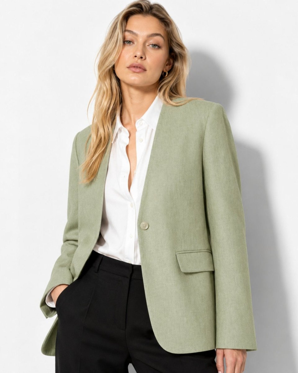 A woman wearing a sage green blazer over a white button-down shirt and black trousers, standing against a plain white wall with a soft shadow.