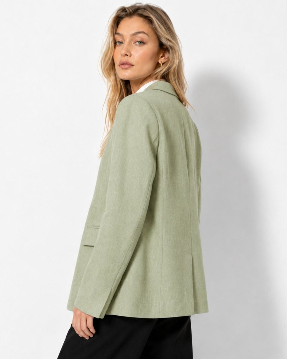 A woman wearing an oversized sage green blazer and black trousers, photographed from a side-back angle against a clean white studio background.