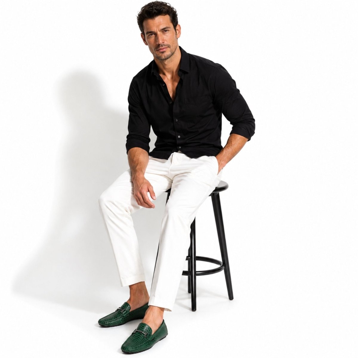A man wearing a black long-sleeve button-up shirt, white trousers, and dark green loafers sits on a black metal stool against a plain white studio background.