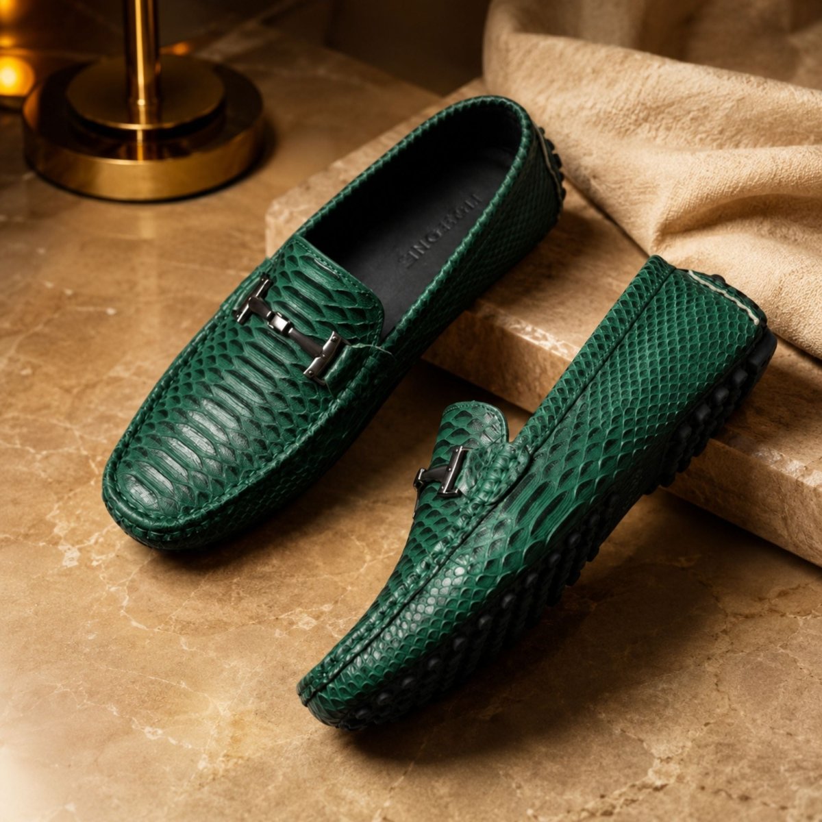 A pair of green snakeskin-textured leather loafers with black metal accents, displayed on a beige stone surface next to a draped beige fabric.