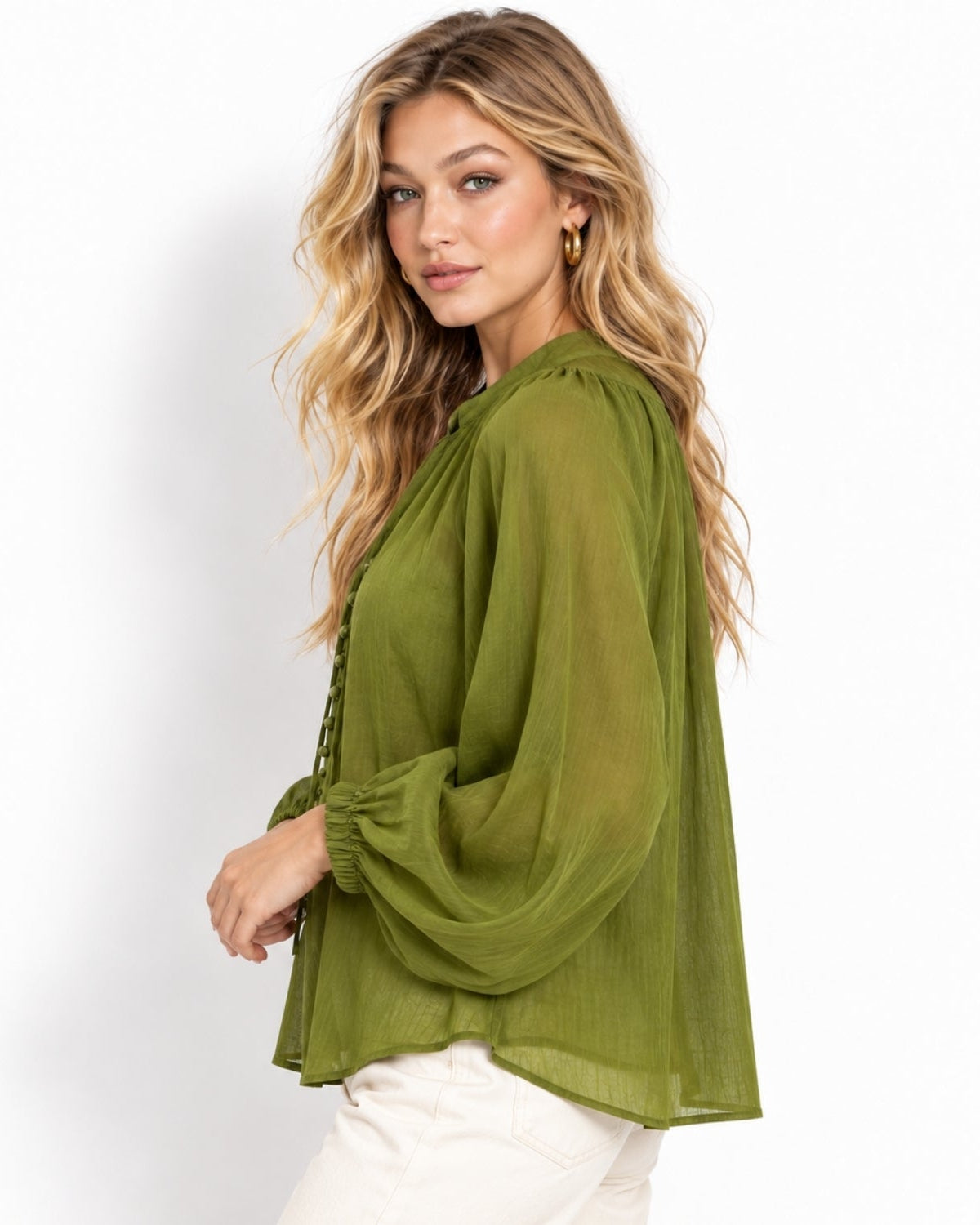 A woman wearing a semi-sheer olive green long-sleeve blouse with gathered cuffs and a button-up front, paired with white pants, photographed against a clean white studio background.