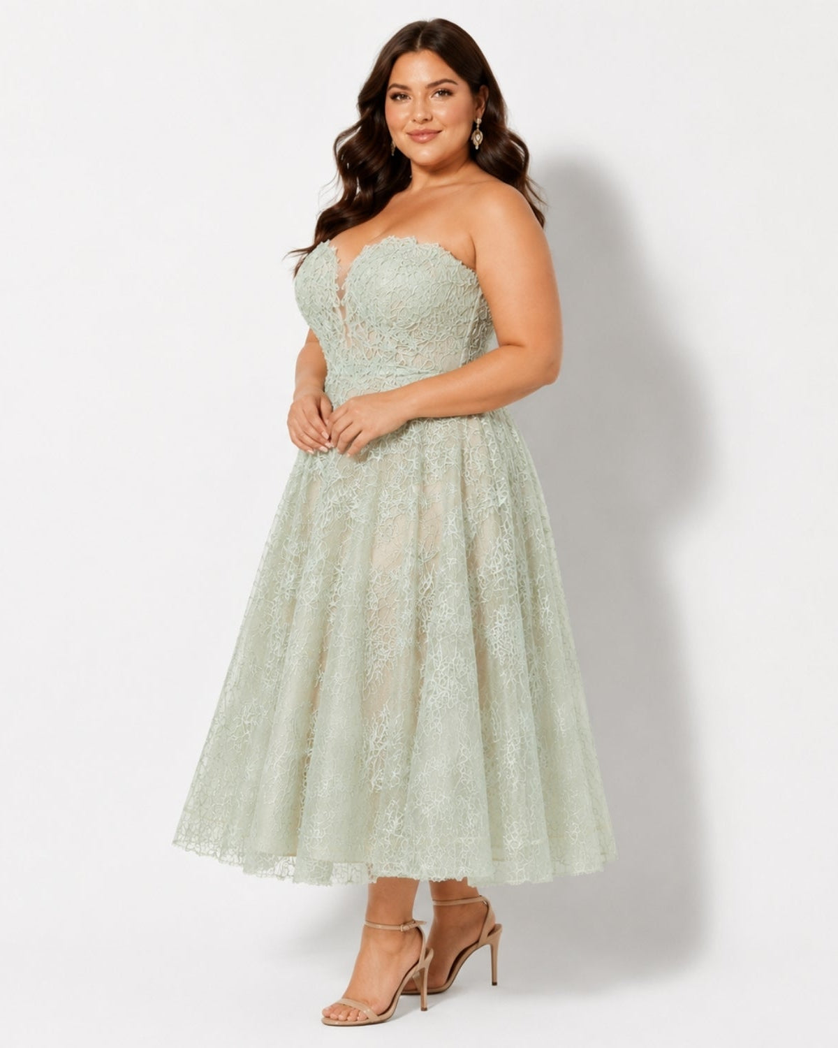 A woman models a strapless light sage green lace midi dress with a sweetheart neckline and a flared skirt, paired with nude high-heeled sandals against a plain white studio background.