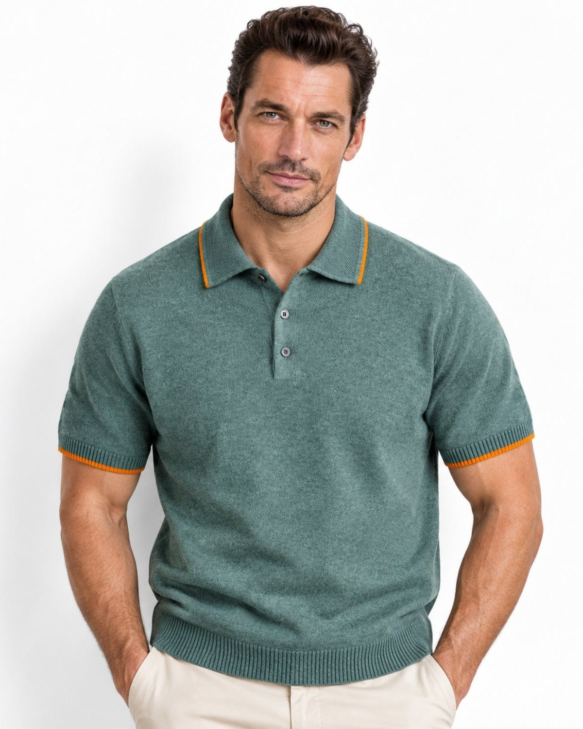 A man wearing a short-sleeve knit polo shirt in muted teal green with thin orange contrast trim at the collar and sleeve cuffs, paired with light beige pants, against a clean white studio background.