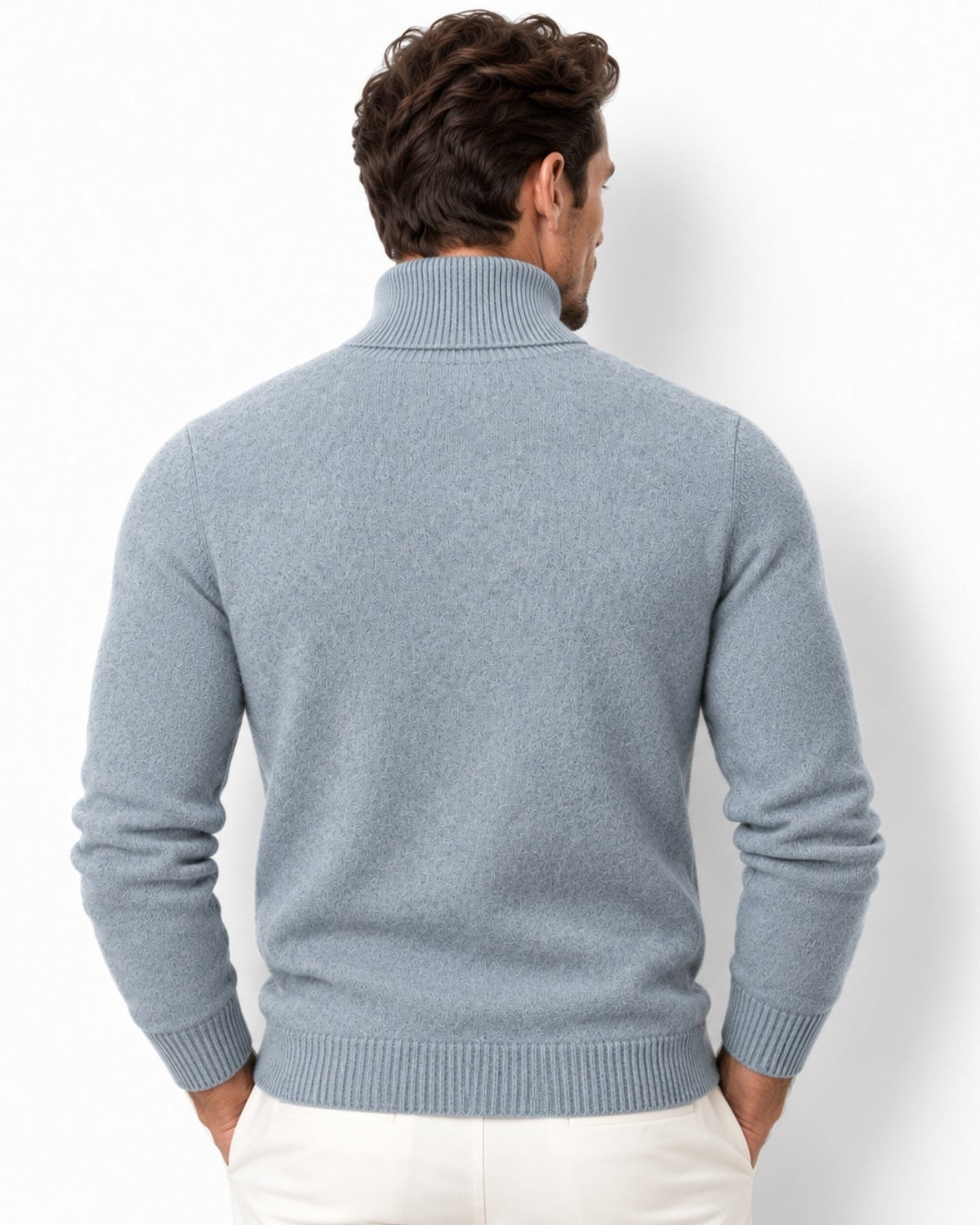 A man viewed from the back wearing a green ribbed turtleneck sweater and off-white trousers, standing against a plain white studio background.