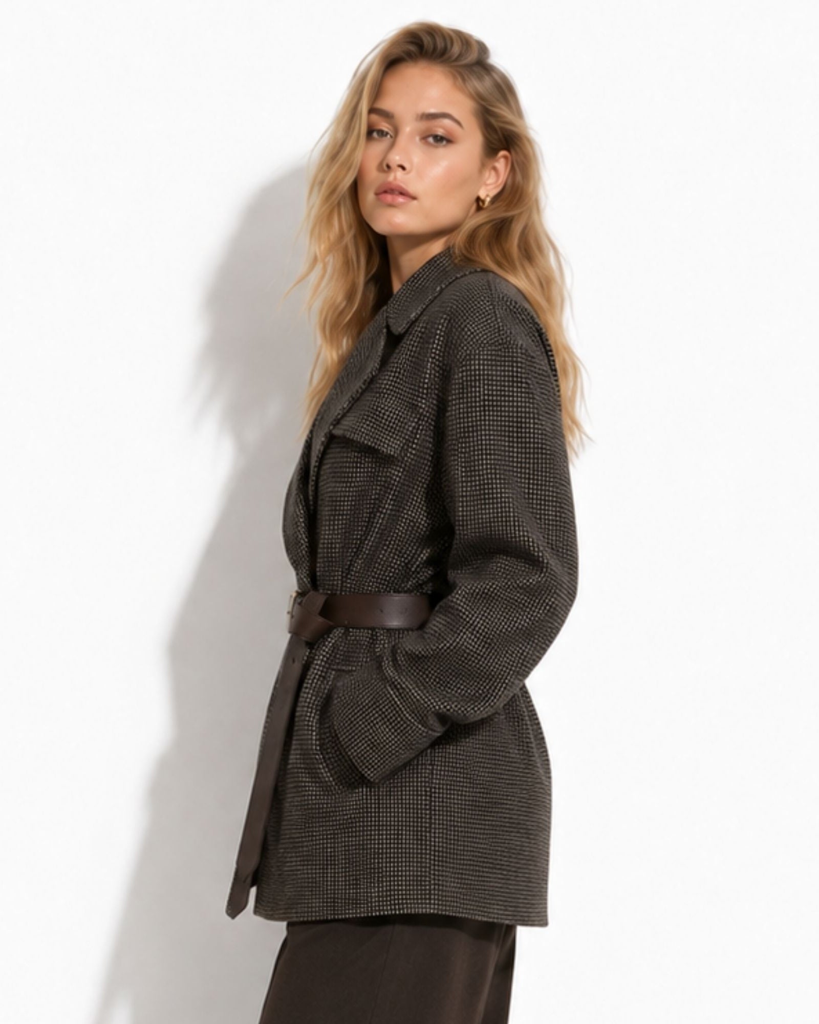 A woman wearing an oversized dark brown and black houndstooth belted coat, posed against a white wall with a soft shadow.