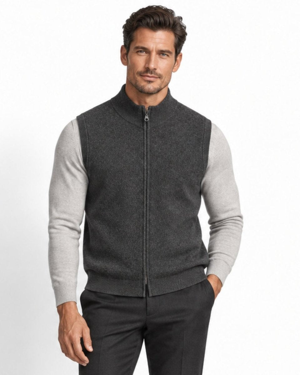Handsome man wearing a grey textured full zip sweater vest over a light grey long sleeve knit, standing with hands in pockets against a white studio background.