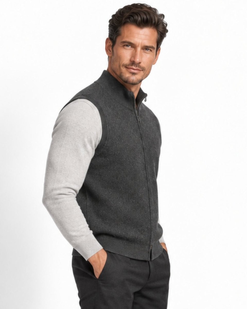 A man wearing a gray zip-up knit vest over a light gray long-sleeve shirt and dark pants, standing against a plain white studio background.