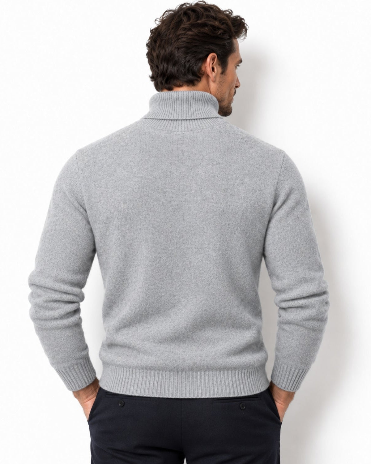 A man viewed from the back wearing a light gray ribbed turtleneck sweater and dark trousers, standing against a plain white studio background.