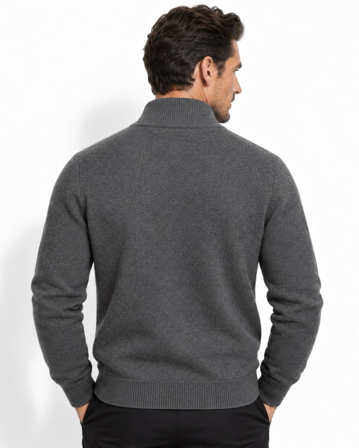 A man viewed from the back wearing a dark gray ribbed mock neck sweater and black trousers, standing against a plain white studio background.