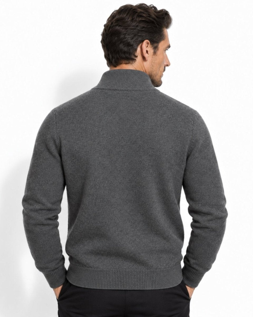 A man viewed from the back wearing a dark gray ribbed mock neck sweater and black trousers, standing against a plain white studio background.