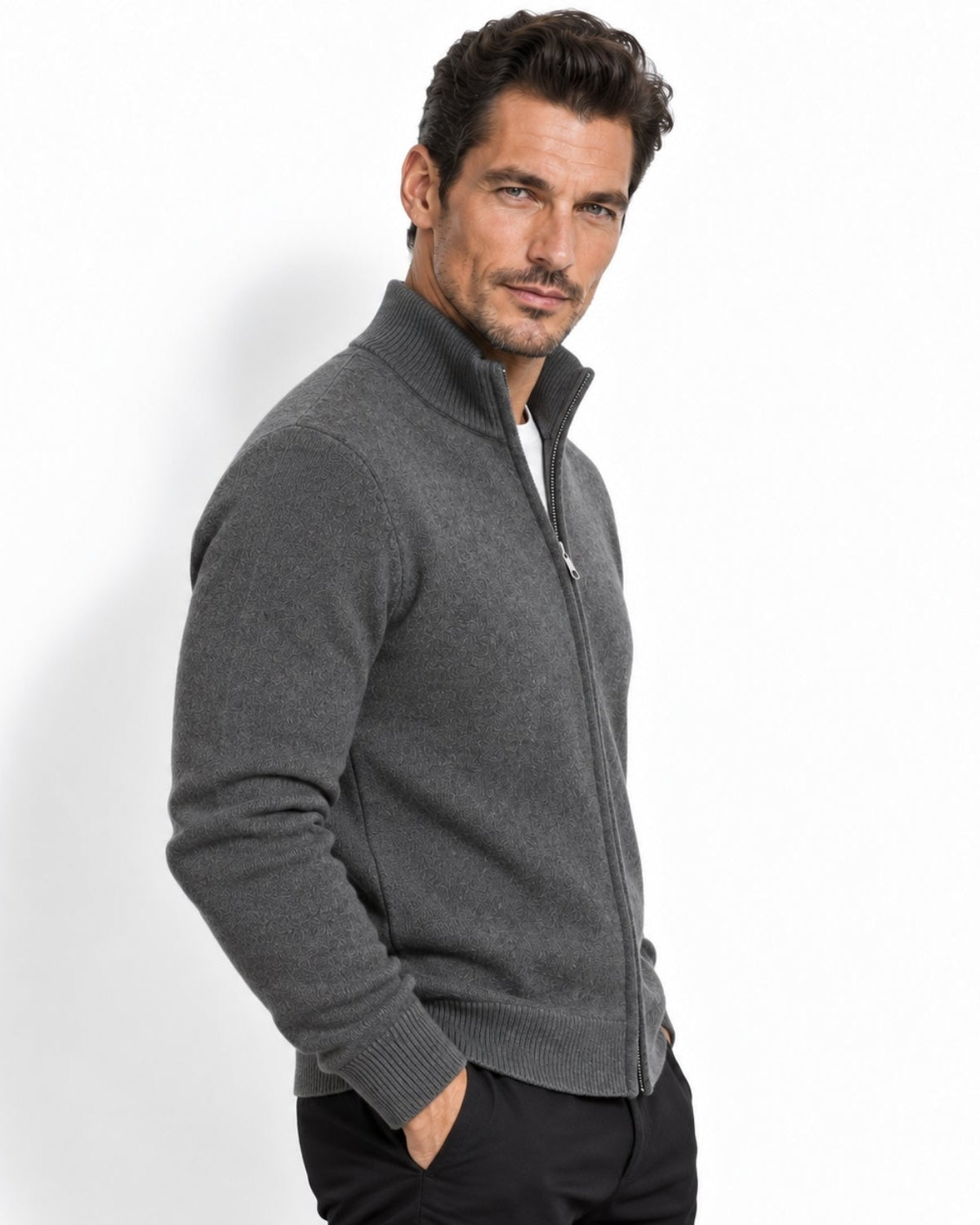 A man wearing a dark gray zip-up knit sweater with a mock neck collar, paired with black trousers, photographed against a clean white studio background.