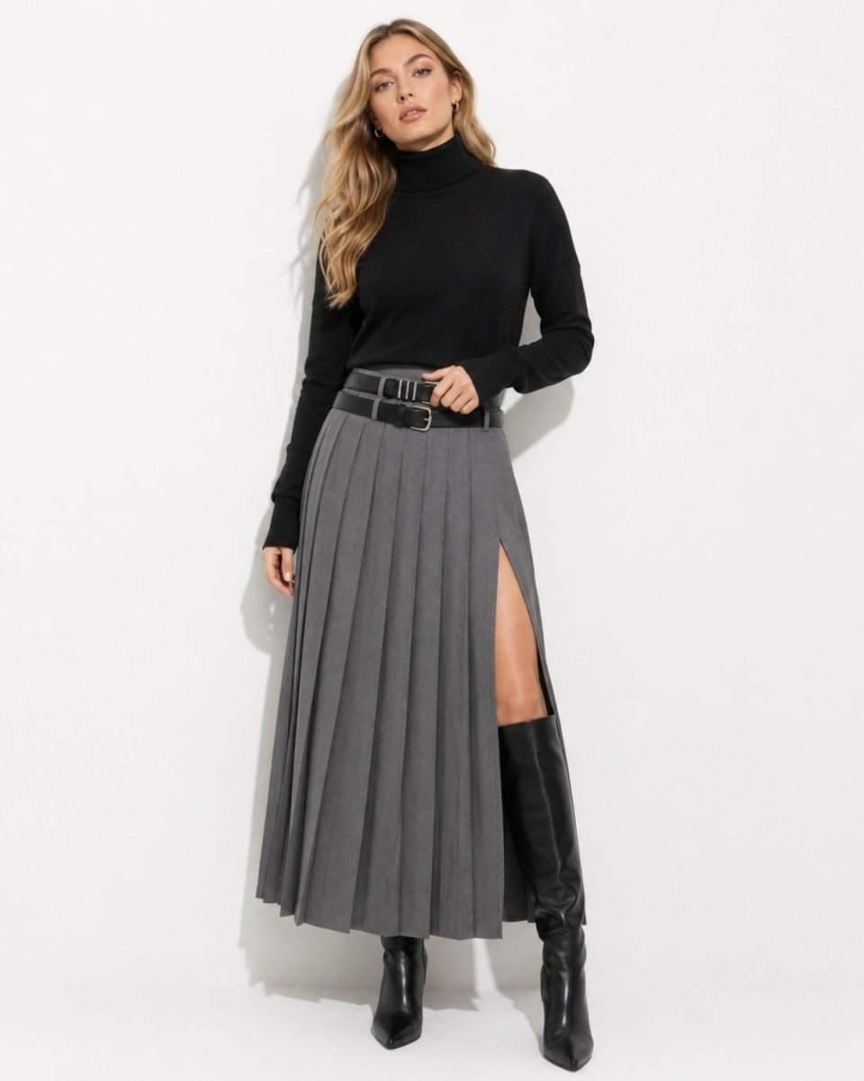 A woman modeling a high-waisted, pleated gray midi skirt with a front slit, styled with a black belt, a black turtleneck, and black knee-high boots against a plain white studio background.