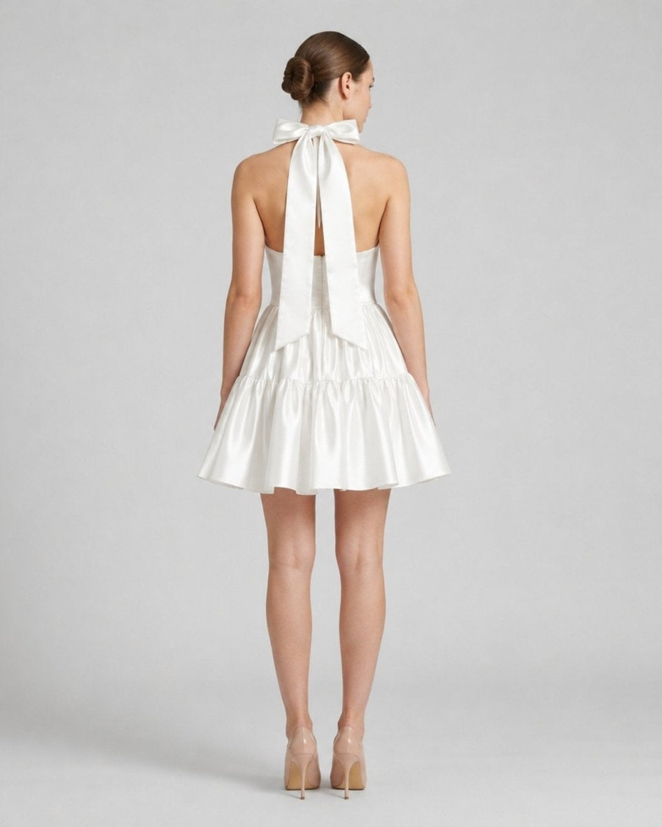 Back view of a white satin open back dress featuring a large tie bow at the neck and a short ruffled skirt.